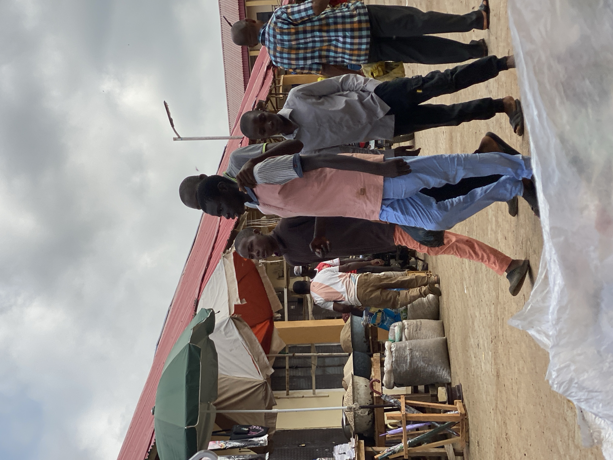The image depicts an outdoor scene in Kafanchan B, Nigeria, likely a marketplace. Several African men are visible, dressed in casual clothing, some in collared shirts and trousers, others in t-shirts and pants. They appear to be walking and interacting with each other. The environment includes buildings with corrugated metal roofs, some with awnings, and a green umbrella. Various items are stored or displayed, possibly for sale, including sacks, containers, and wooden structures. The sky is overcast.