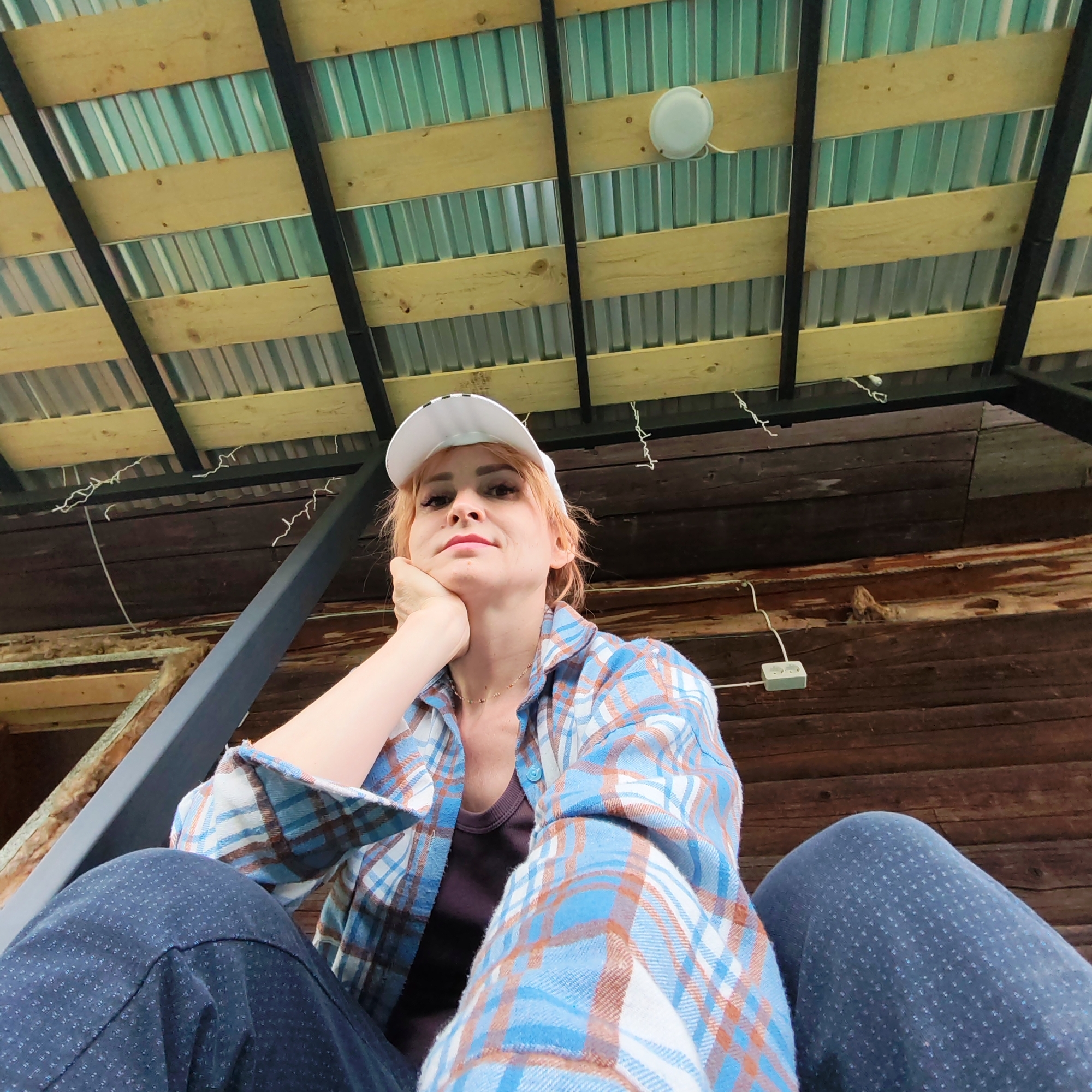 A low-angle shot depicts a woman with reddish-blonde hair wearing a white baseball cap and a blue plaid flannel shirt. She is seated with her legs visible in the foreground, clad in dark blue pants. Her left hand is positioned beneath her chin in a contemplative pose.
Overhead, the structure consists of wooden beams and metal corrugated roofing. A small, circular white light fixture is attached to the wood. An electrical outlet is visible on the wooden wall behind her. Based on the wooden construction, the location appears to be a rustic, possibly outdoor, setting. The city of Chaikovsky, Russia is not discernable from the visual information provided.