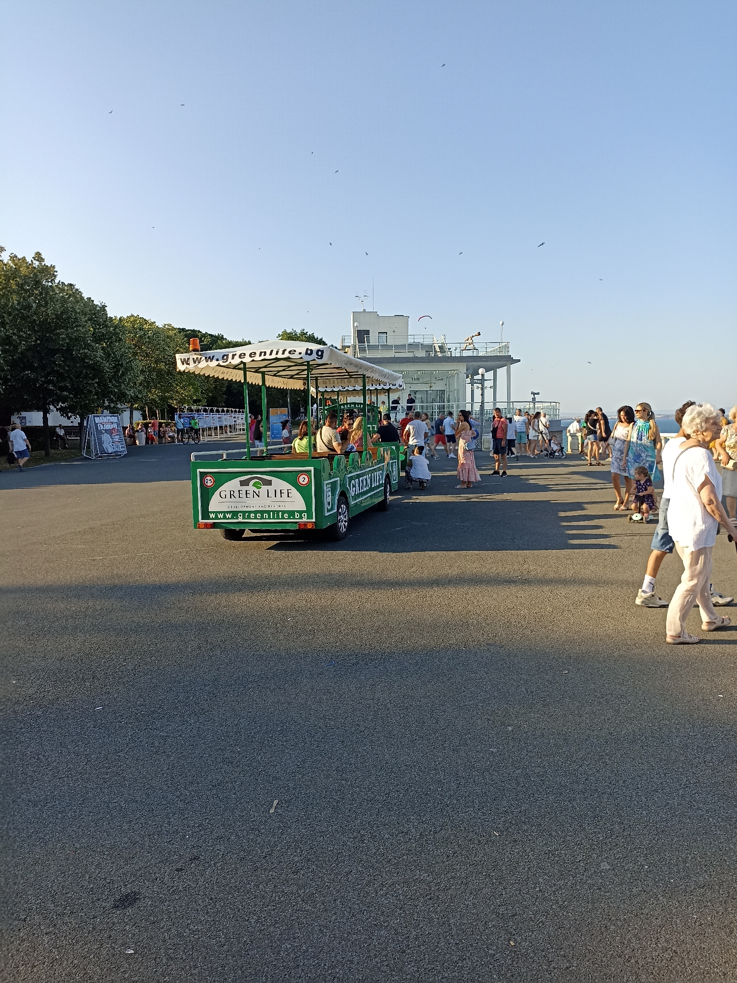 The image depicts a sunny outdoor scene at the Sea Station in Burgas, Bulgaria. In the foreground, an asphalt surface is visible with people walking on it. A green tourist train with a white canopy, labeled "GREEN LIFE" and "WWW.GREENLIFE.BG," is positioned in the middle ground, carrying passengers. A group of people, including adults and children, are seen standing and walking around the train and in the background. Behind the train, a modern building with a balcony and railings is visible, with a clear blue sky overhead. Trees and a large advertising board are also present on the left side of the image. Several birds are flying in the sky. The overall atmosphere suggests a leisurely day with tourists and locals enjoying the waterfront area.