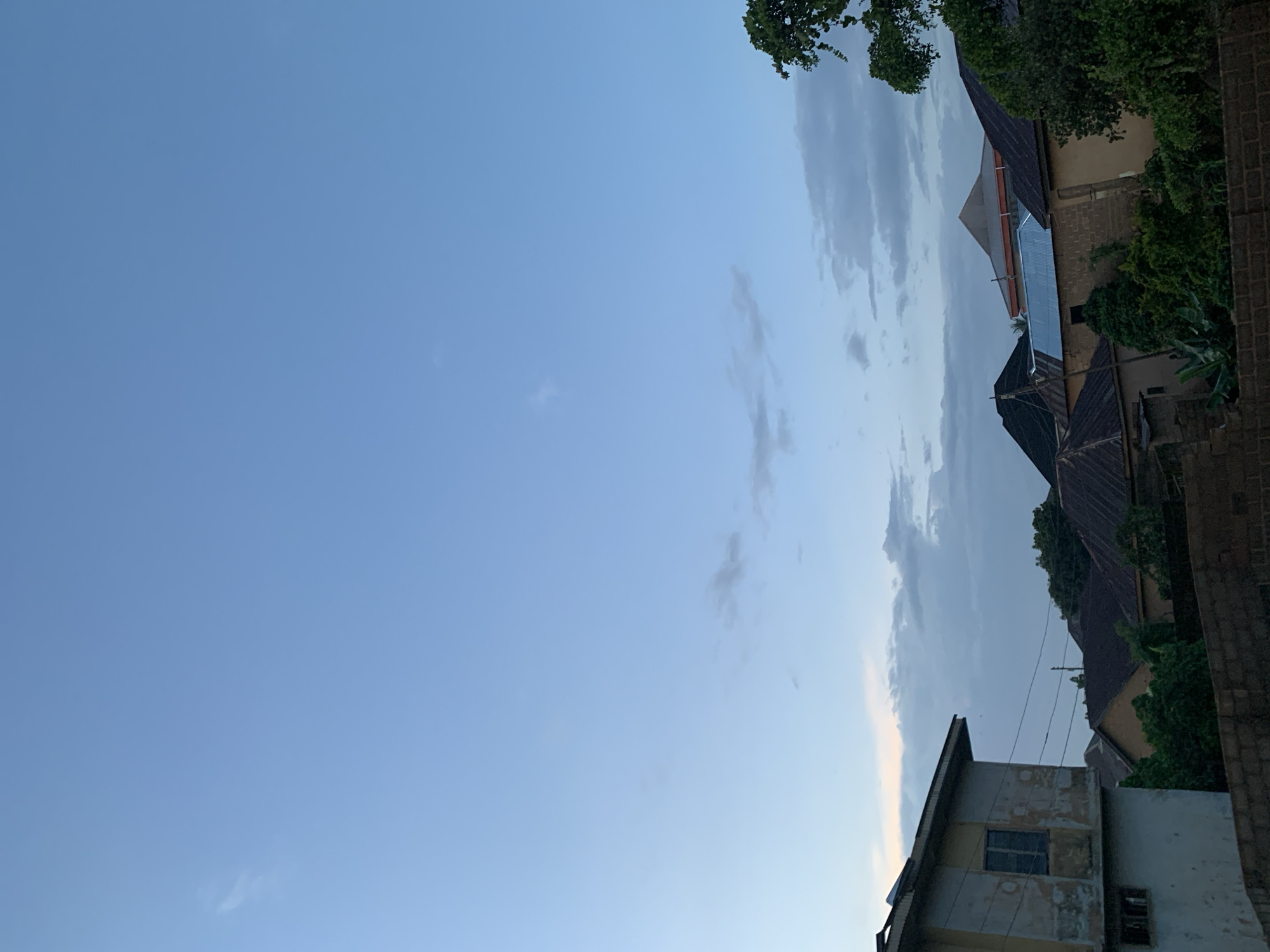 The image depicts a residential area under a pale blue sky with scattered clouds. The foreground is dominated by the side of a weathered, multi-story building with peeling paint, a single window visible on the upper floor, and what appears to be a boarded-up window on the lower level. Beyond this building, the roofs of several other houses are visible, characterized by corrugated metal and tiled materials in various shades of brown and gray. Lush green trees and foliage are interspersed between the buildings, suggesting a tropical or subtropical climate. The overall impression is one of a densely populated urban or semi-urban environment, possibly at dusk or dawn given the soft lighting and subdued colors of the sky.
