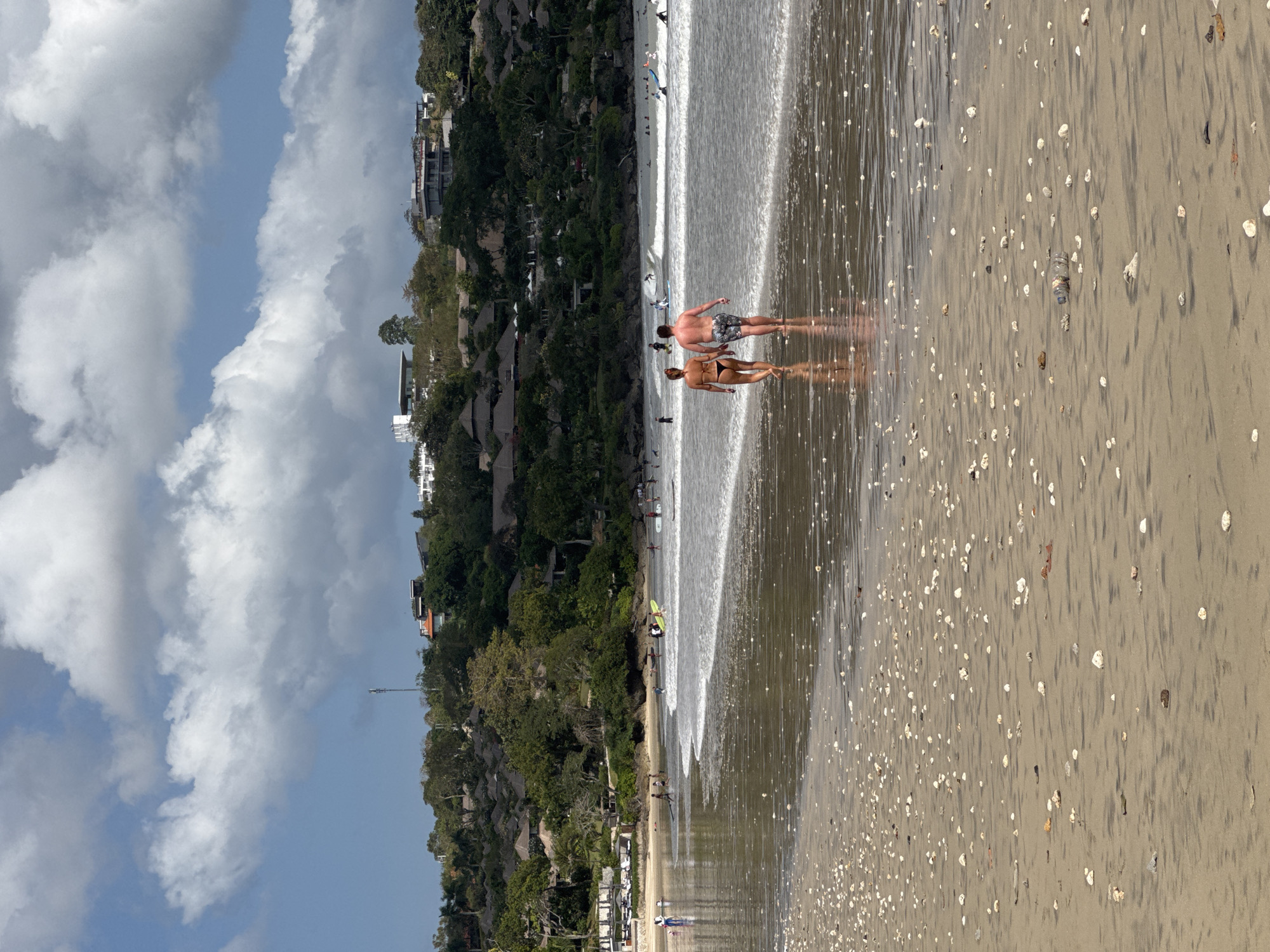 The image shows a wide shot of Kuta Selatan beach in Indonesia. A couple is walking away from the camera on the wet sand, with the ocean on their left. The man is wearing patterned swim shorts, and the woman is wearing a black bikini. The ocean has small waves and is populated with several surfers. In the background, a lush green hill is covered with trees and buildings. The sky is blue with fluffy white clouds. The foreground is filled with sand and scattered seashells, with a plastic water bottle lying on the sand.