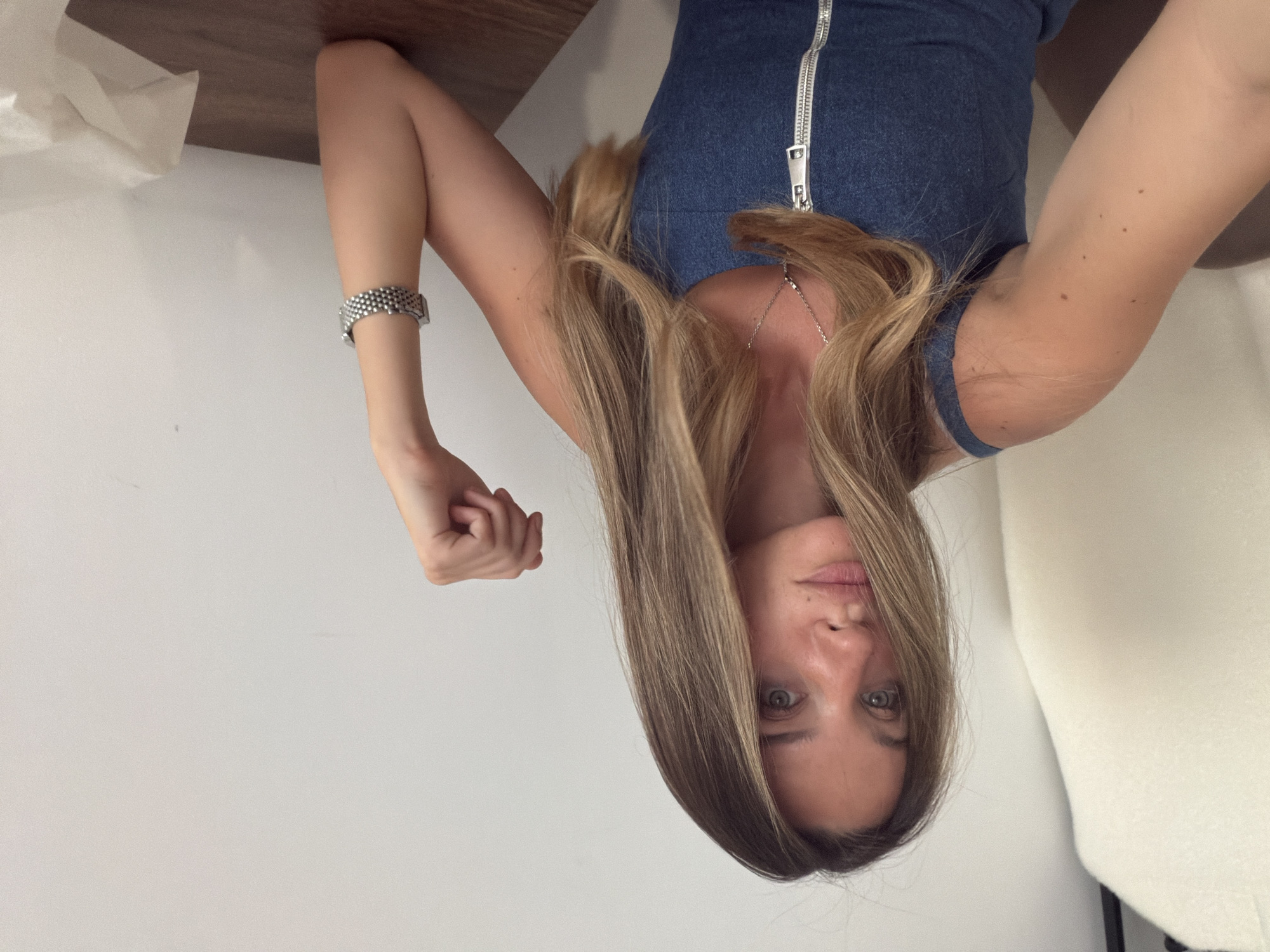 A woman with long, light brown hair is wearing a blue denim dress with a silver zipper down the front. She is also wearing a silver necklace and a silver watch on her left wrist. The background is a white wall and a light brown surface.
