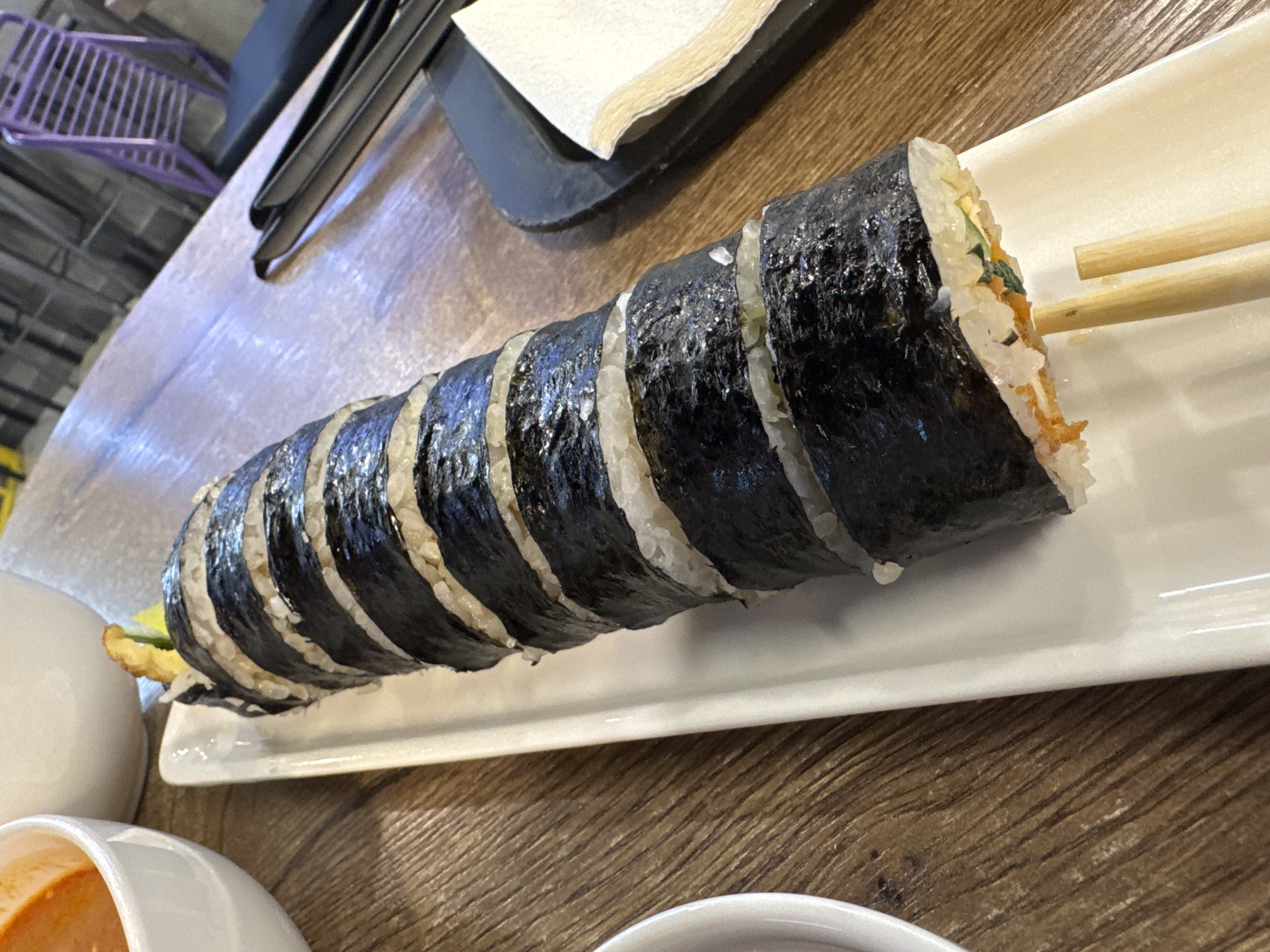 The image depicts a plate of sliced kimbap, a Korean seaweed rice roll, resting on a white rectangular plate on a wooden table. Chopsticks are placed beside the kimbap. Below the plate are the edges of two white bowls. The scene is indoors, likely in a restaurant. Beyond the table are hints of a metallic, industrial-looking structure, potentially shelving or chairs. The location in Mozhvai, Russia cannot be verified with the provided image and information.