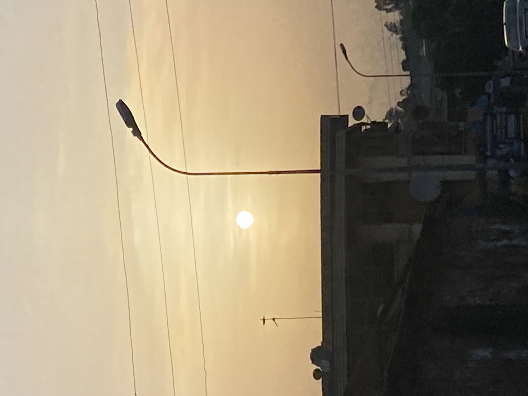 The image shows a landscape in Abofifia, Nigeria, at sunset. The sun is visible in the center of the frame, with the sky appearing in shades of orange and yellow. A street light is positioned to the left of the sun, with power lines visible in the sky. Buildings and trees are seen on the right side of the image. There are no people visible.