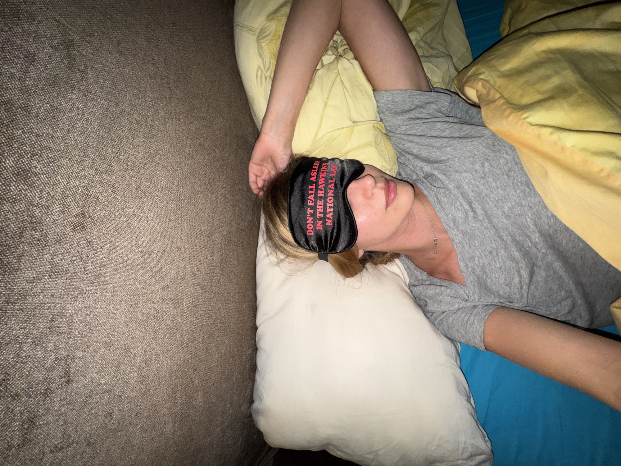 A woman is lying down, wearing a sleep mask that says "DON'T FALL ASLEEP IN THE HAWKING NATIONAL LAB." She is lying on a white pillow, a yellow pillow, and a blue sheet. The location is unknown.
