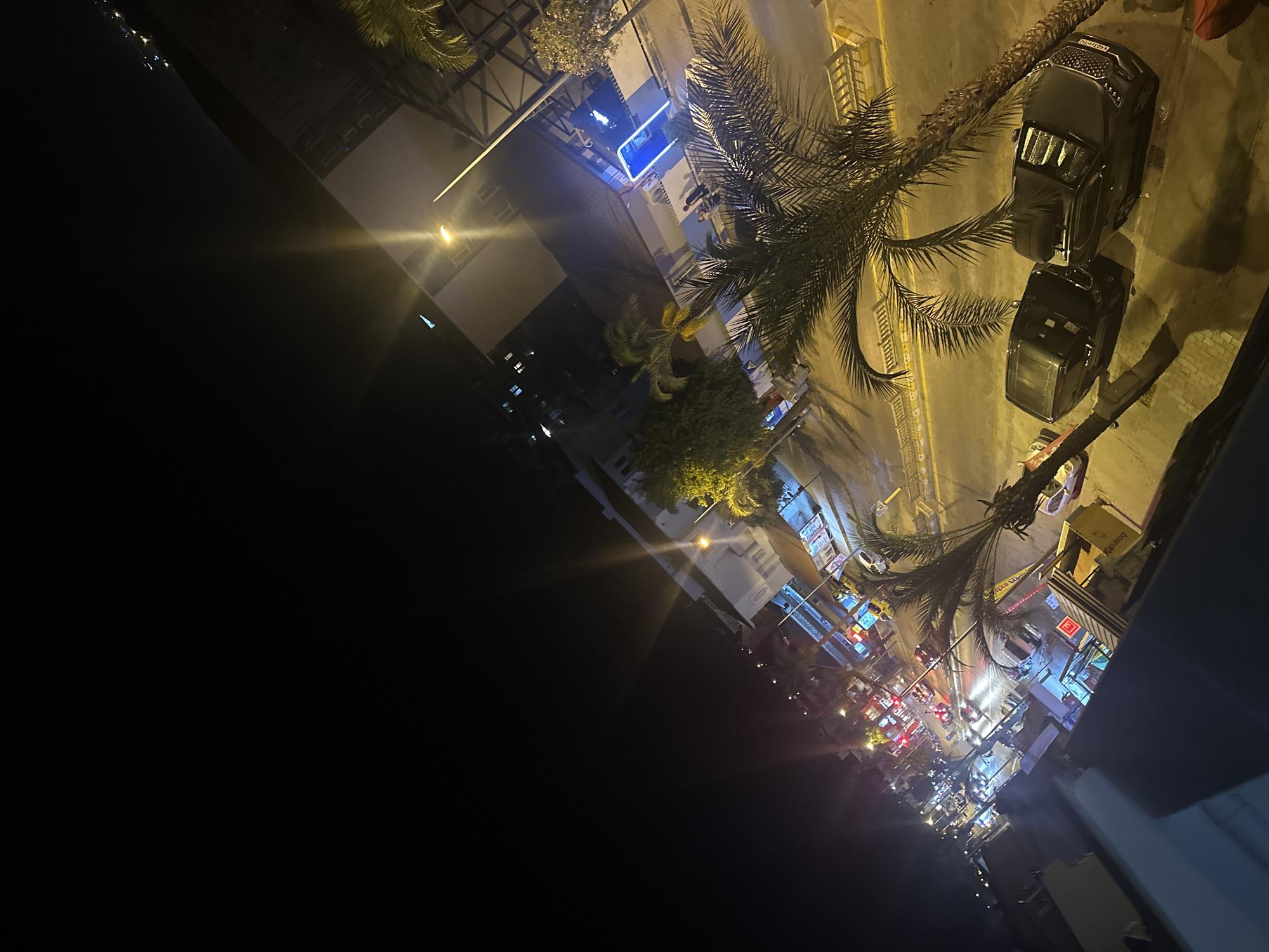 The image depicts a nighttime street scene in Fethiye, Turkey. The perspective is from an elevated viewpoint, looking down onto a street lined with buildings and palm trees. Several vehicles, including at least two dark-colored SUVs parked on the side of the road, are visible. Pedestrians are present on the sidewalks and some are interacting with each other. The street is illuminated by streetlights and the colorful neon signs of businesses, suggesting a commercial or entertainment district. Buildings along the street have multiple stories and balconies. The sky is dark, indicating it is well after sunset, and distant lights suggest further development or the presence of hills or mountains in the background. The overall impression is of a vibrant, active urban environment at night.