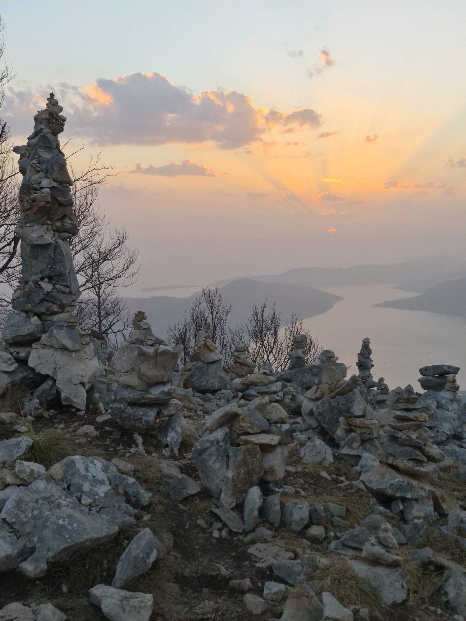 The image depicts a mountainous landscape at sunset, with a body of water visible in the distance. The foreground is dominated by numerous cairns, or stacked stones, of varying sizes. These cairns are artfully arranged on a rocky terrain, suggesting a hiking trail or a sacred site. In the background, the sky is painted with soft hues of orange, pink, and blue, indicative of the setting sun, with rays of light emanating from behind the clouds. The overall scene conveys a sense of tranquility and natural beauty, typical of elevated viewpoints in regions like Kuk, Montenegro, which is known for its stunning natural landscapes and opportunities for hiking and exploration.