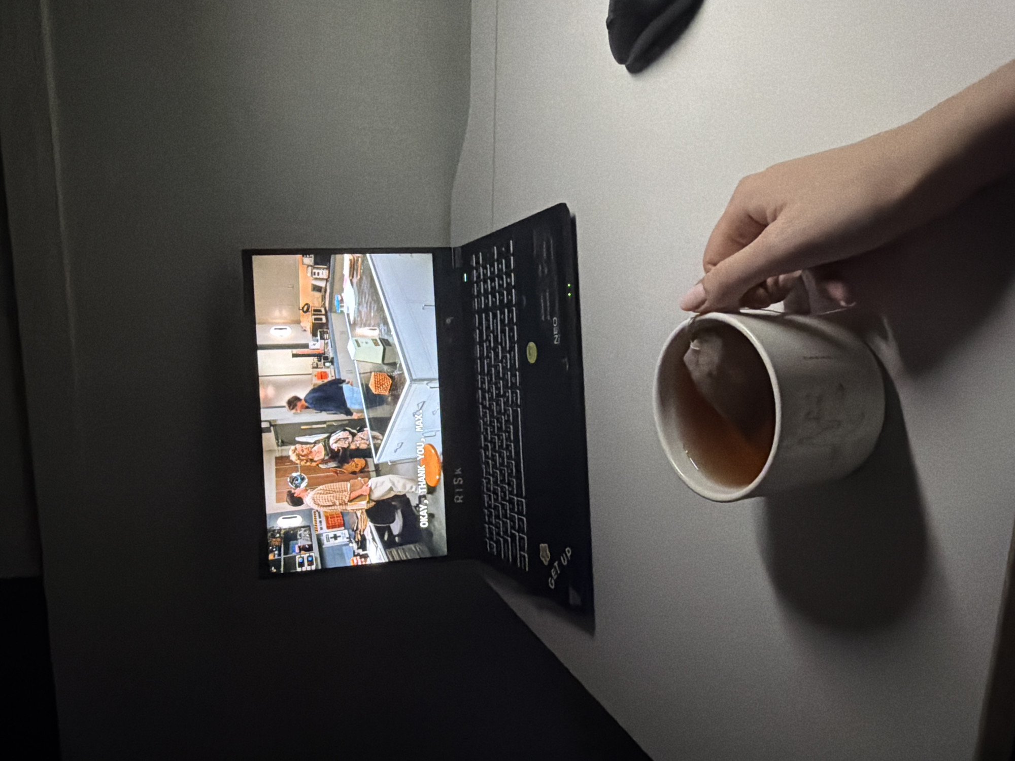 The image captures a dimly lit scene, likely indoors, on a desk in Dongducheon-si, South Korea. A laptop, displaying a scene from a television show or movie, is open. The show appears to be set in a kitchen, with three individuals visible on screen. Below the screen, text reads "OKAY, THANK YOU, MAX." The laptop itself has a sticker on the left side that says "GET UP" and the brand "RISK" is visible on the palm rest. Two green indicator lights are illuminated on the laptop's keyboard area. In the foreground, a white mug filled with tea is held by a person's hand, which has neatly manicured nails. The hand is positioned to hold the mug. A dark object, possibly a computer mouse, is located to the right of the mug. The overall environment suggests a personal space, perhaps a bedroom or study, for relaxation and entertainment.
