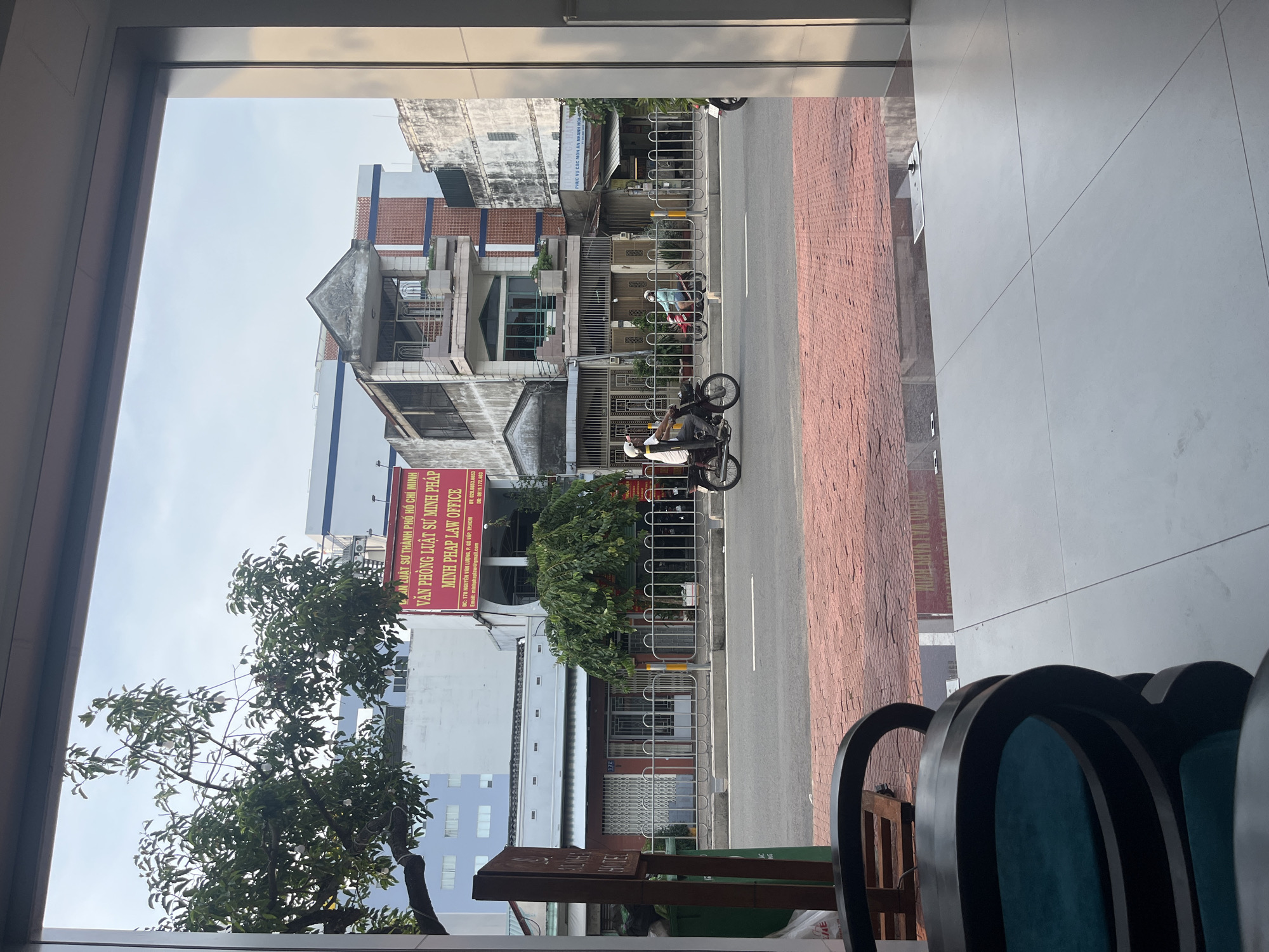 The image shows a view of a street scene from indoors, looking through a large window. In the foreground, a portion of a dark-colored chair with teal upholstery is visible on the left. The outdoor scene depicts a street with red brick paving in the foreground and asphalt further back. Several buildings line the street, featuring a mix of architectural styles. A red and white sign with Vietnamese text is mounted on one of the buildings. Two people on motorcycles are traveling down the street. A tree with green foliage is prominent on the left side of the street. The sky is overcast.