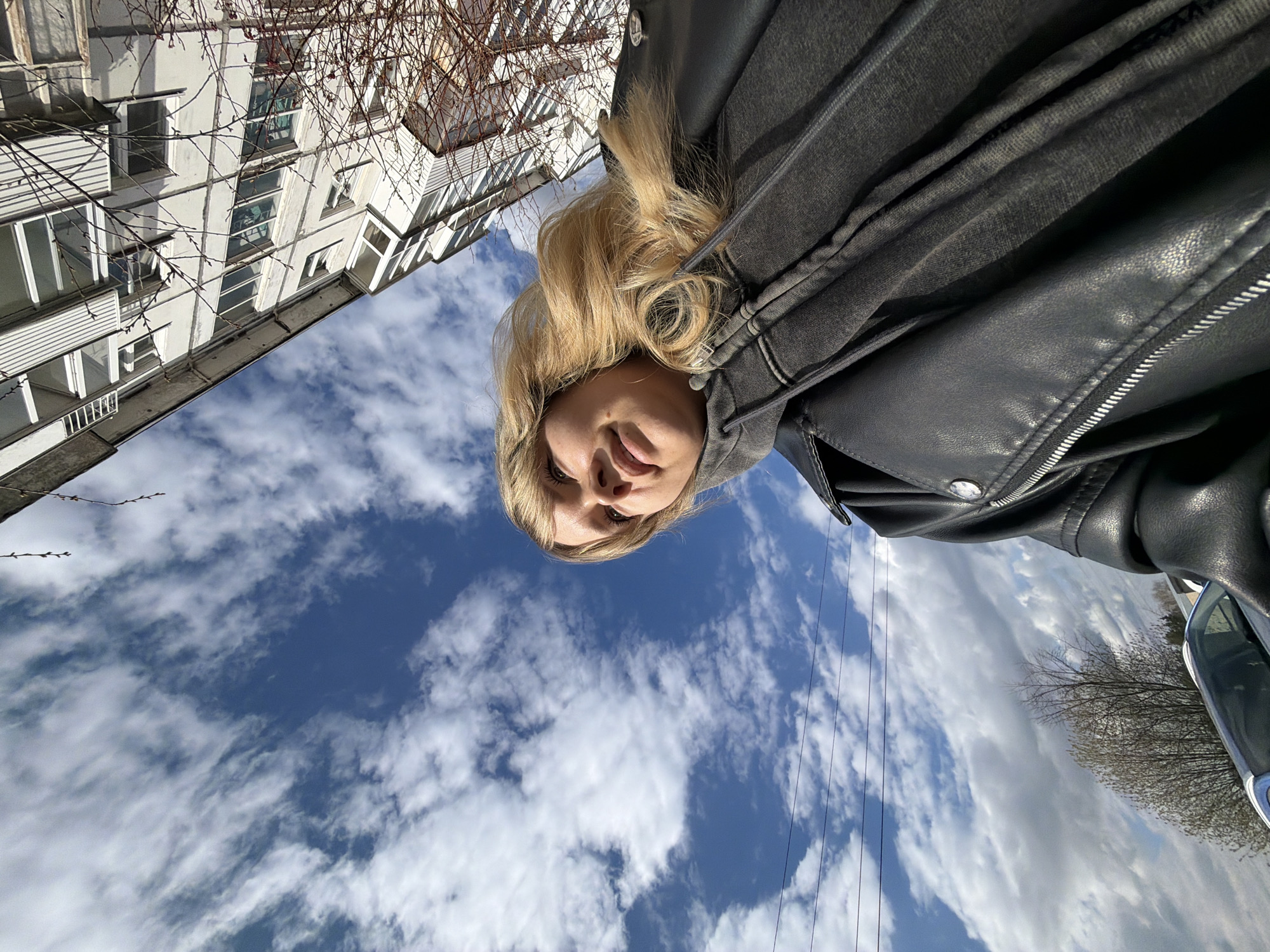 The image is a low-angle selfie taken outdoors in Chaikovsky, Russia. A fair-skinned blonde woman with shoulder-length curly hair and wearing a grey hooded sweatshirt under a black leather jacket is centrally framed against a blue sky with scattered white clouds. Above her head and slightly to the left is the corner of a white building with multiple windows. Bare tree branches are partially visible in the upper left corner. Power lines run diagonally across the sky. A tree with budding leaves and part of a silver vehicle are visible in the lower right corner.