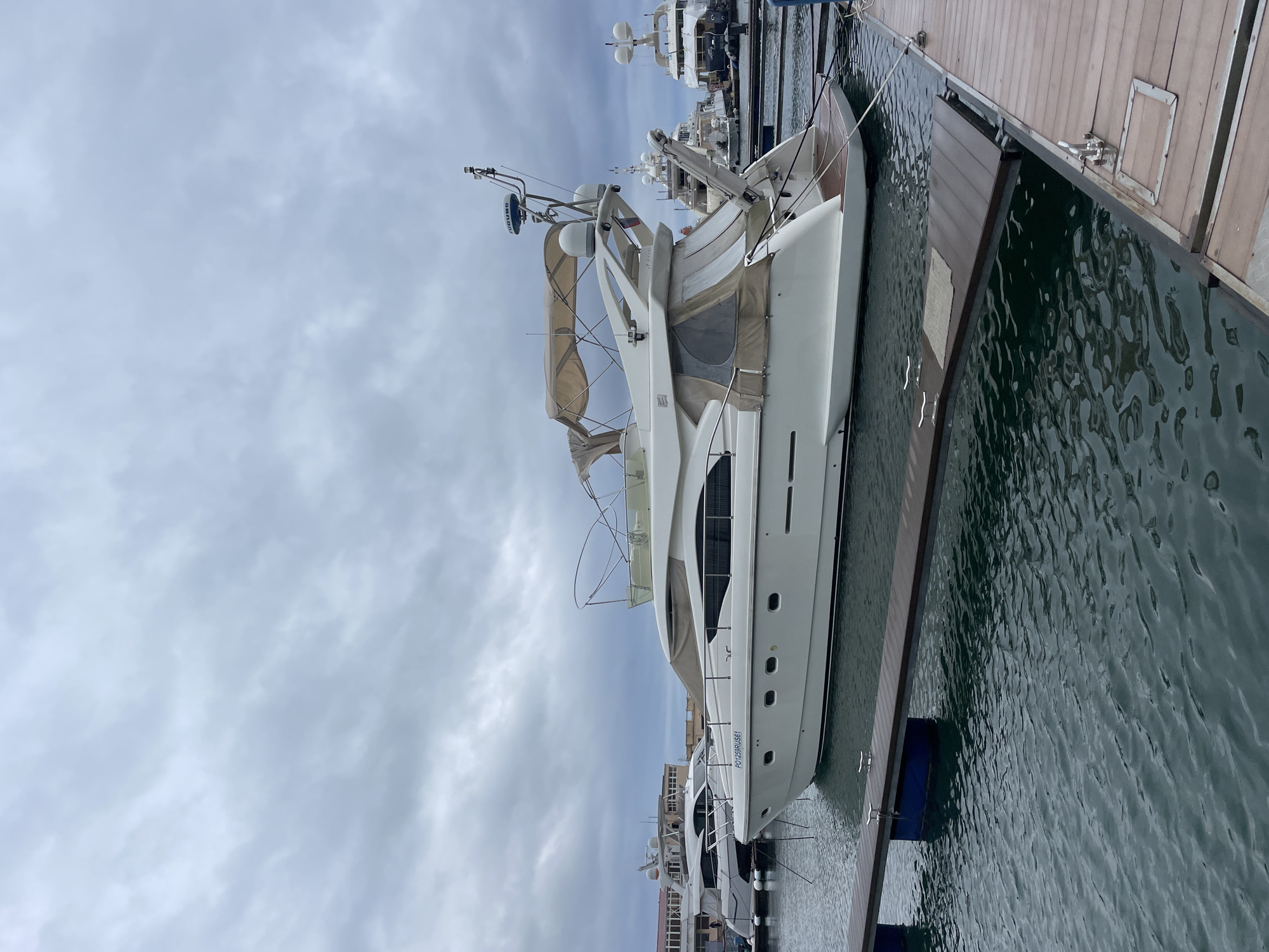 A horizontal, full shot captures a white, medium-sized yacht docked beside a brown wooden pier. The yacht is equipped with a radar dome and a beige canvas awning, and is tethered to the pier. The murky green water surrounds the yacht and pier, and a series of other yachts can be seen in the background, along with low-rise buildings. Above, a cloudy sky fills the frame. The visible license plate reads: PO 1259 RU 561. The image appears to be taken in Sochi, Russia.