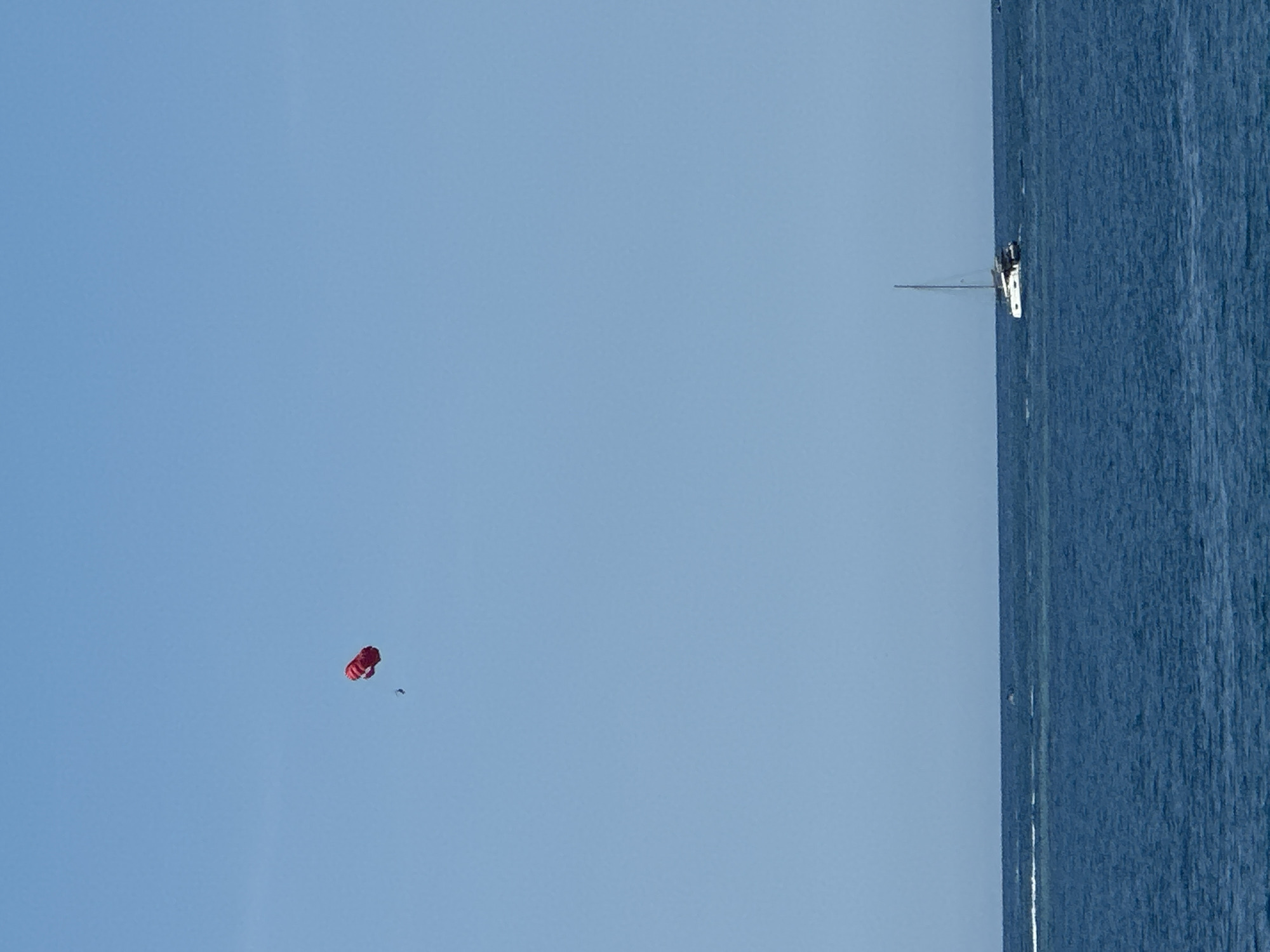 The image presents a wide outdoor scene featuring a clear, light blue sky dominating the upper two-thirds of the frame, set above a deep blue sea occupying the lower third. In the upper left quadrant of the sky, a single person is visible, suspended in mid-air beneath a deployed, bright red parasail. On the surface of the water in the lower right quadrant, a white catamaran with a single mast is actively sailing. A faint white wake is discernible on the far left edge of the water, indicating motion. The scene is located in Budva, Montenegro.