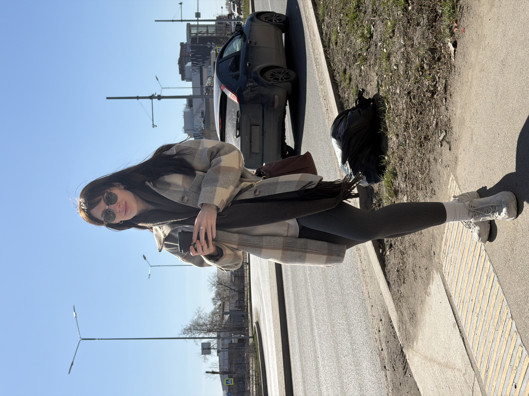 A young woman with long, dark hair stands on a sidewalk next to a street. She is wearing sunglasses, a light-colored, plaid overcoat, gray leggings, white socks, and white sneakers. She is holding a black object in her hands. A dark brown handbag hangs on her shoulder. Beside her, a black trash bag rests on a patch of dirt and sparse vegetation. Behind the woman, a black car is parked along the curb of the street. Street lights and buildings are visible in the background under a clear, blue sky. The image appears to be taken in the city of Perm, Russia.