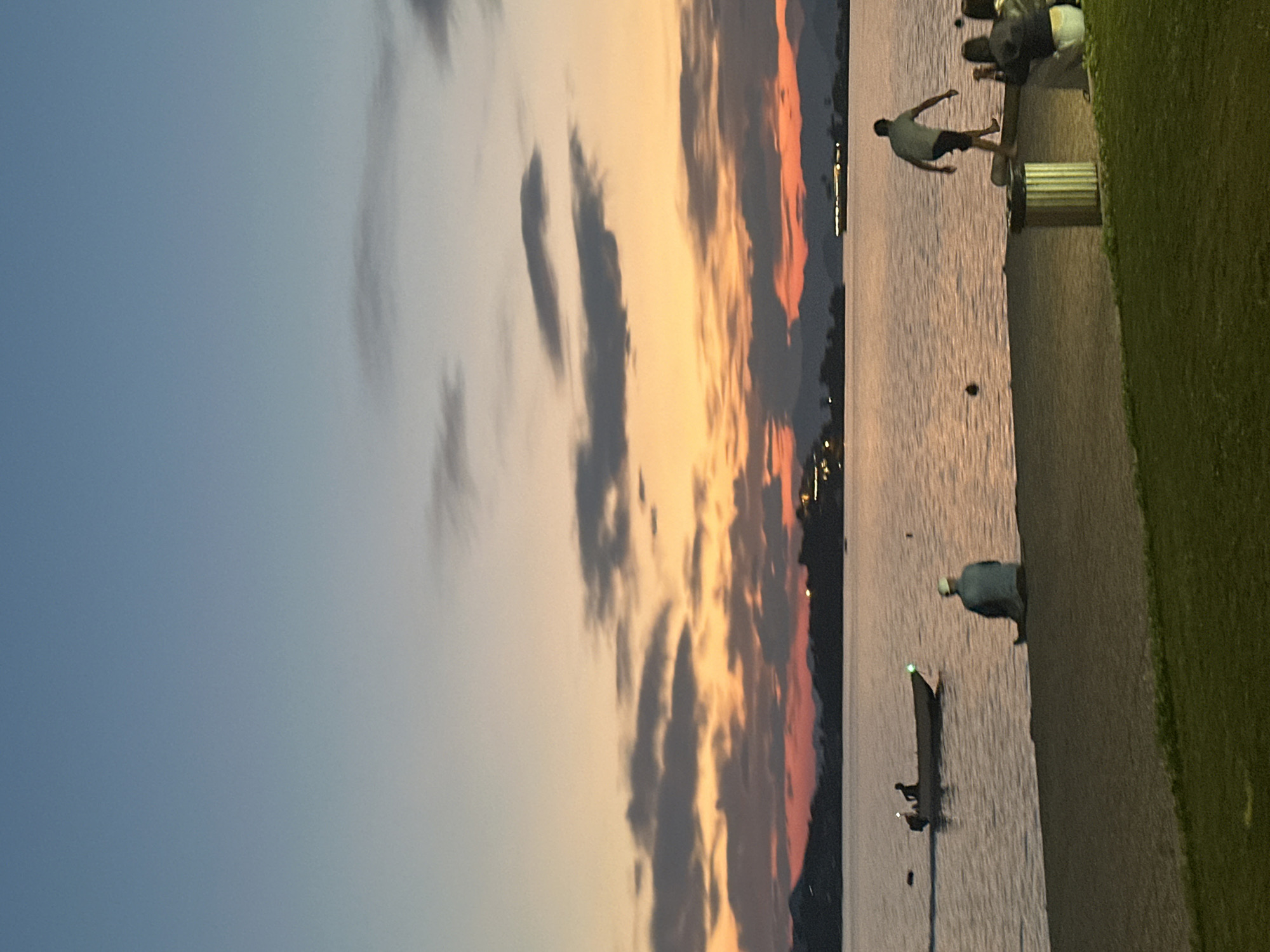 The image captures a sunset scene at Fethiye, Turkey. The sky is a blend of orange, pink, and grey clouds, transitioning to blue. Silhouetted against the sunset is the coastline and possibly mountains in the distance. In the foreground, there is a grassy area on the right, with several people visible. One person is in mid-air, poised as if about to jump into the water. Farther down, there is a boat with two people in it, and another person standing near the water's edge. The water surface reflects the sunset colors.