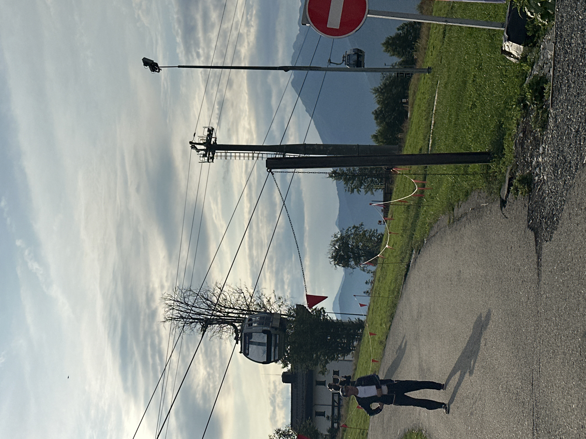 A man stands on a paved path in the foreground, facing left. He is wearing a dark jacket with blue stripes, dark pants, and sneakers. The path curves to the left and up a grassy slope dotted with red flags. In the background, a cable car ascends a mountain. Further back, a building with a dark roof is visible, with trees surrounding it. Mountains stretch across the horizon under a cloudy sky. To the right, a traffic sign with a red circle and a white horizontal bar is visible, indicating a prohibition. The scene appears to be located in Estosadok, Russia, likely at a ski resort or similar mountain recreational area.