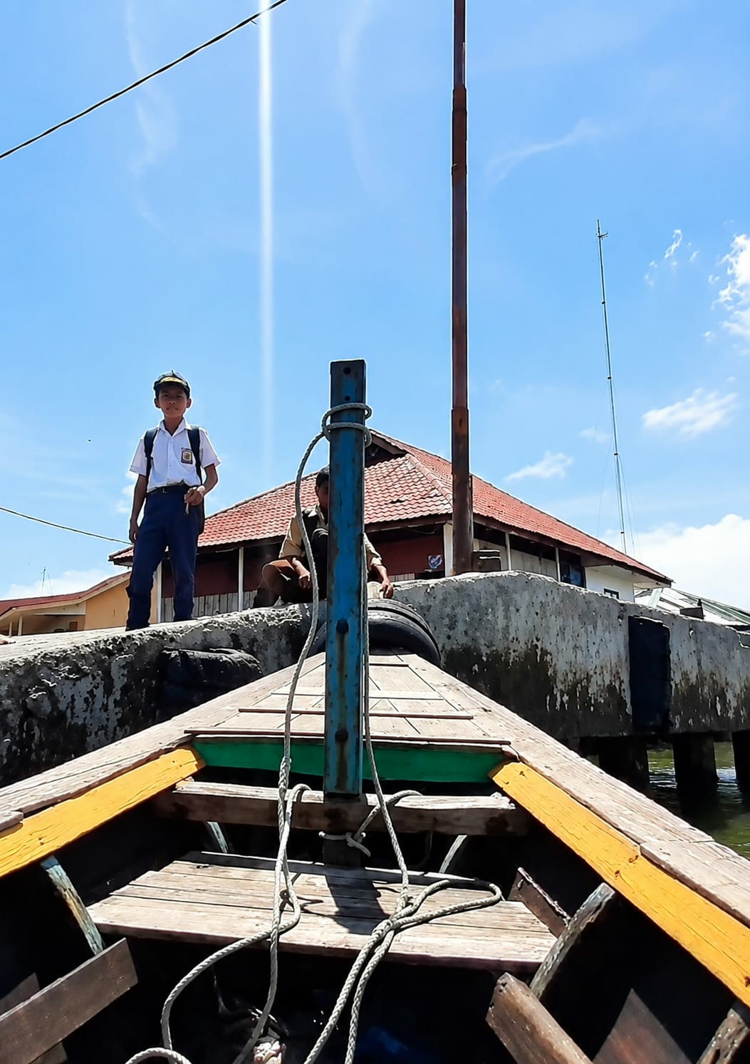 A low-angle shot shows the wooden bow of a boat, with green and yellow accents, facing a concrete pier under a bright blue sky with scattered clouds. A young boy in a school uniform, white shirt, dark pants, and a cap, stands on the pier to the left, looking towards the camera with a backpack on his shoulders. Behind him, a man in a tan shirt and dark vest is partially visible, sitting on the pier. A tall, rusty red pole is on the right side of the pier, and a thinner green pole is visible further in the background. Buildings with red tiled roofs are situated behind the pier. The scene suggests a sunny day, likely in a coastal or riverine area, possibly a transportation hub for students. The specific location in the City of Medan, Indonesia, is not visually identifiable from the image alone, but the presence of school children and water transportation is common in various parts of the city.