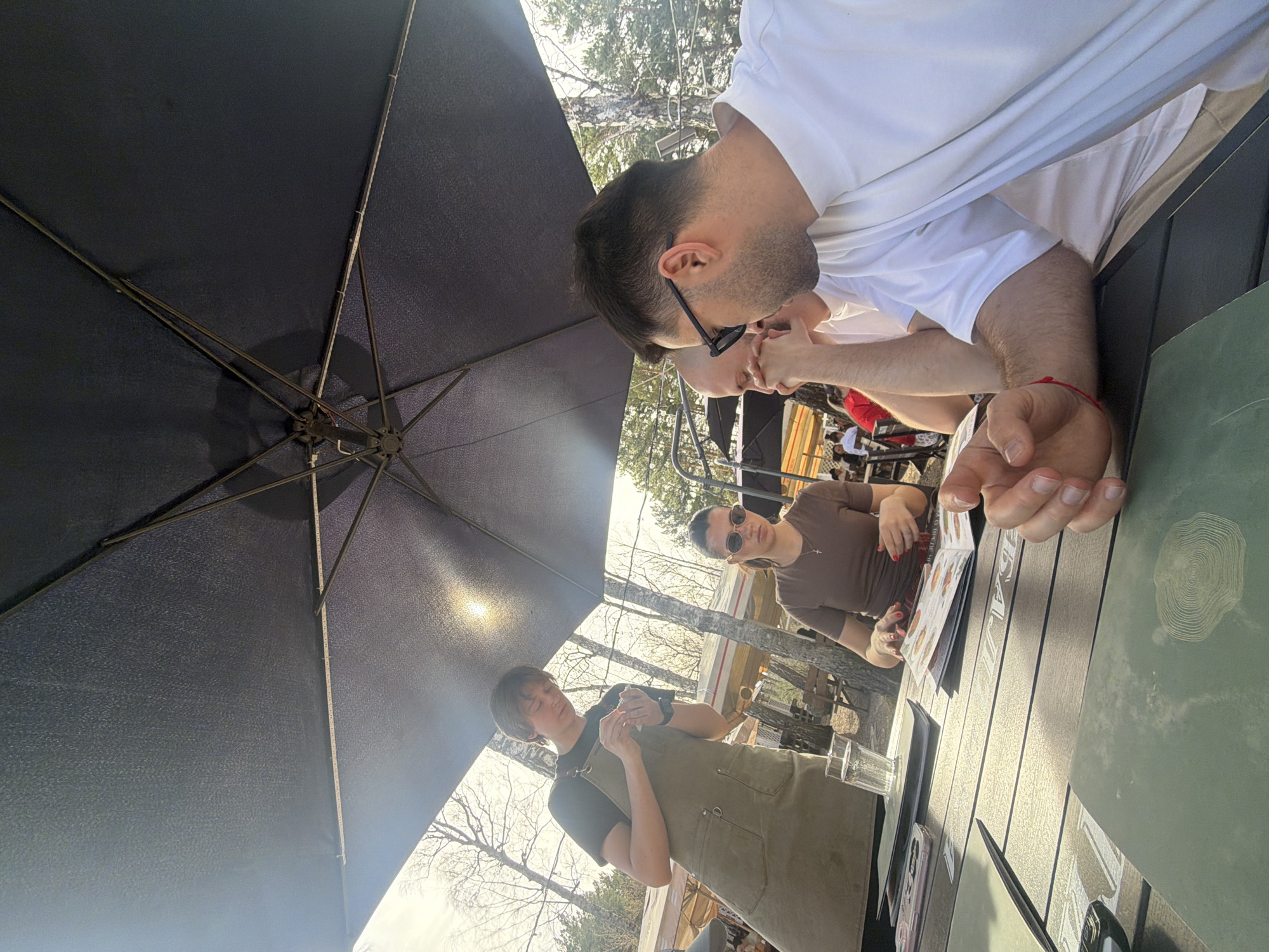 The image depicts a restaurant scene, likely outdoors given the presence of an umbrella, trees, and daylight. A man in a white shirt is seated at the table with another person partially visible near him. A woman wearing sunglasses and a brown shirt is visible across the table, looking at a menu. A server wearing an apron is interacting with the table. The table has menus, glasses, and other restaurant items. The location is Krasnoyarsk, Russia.
