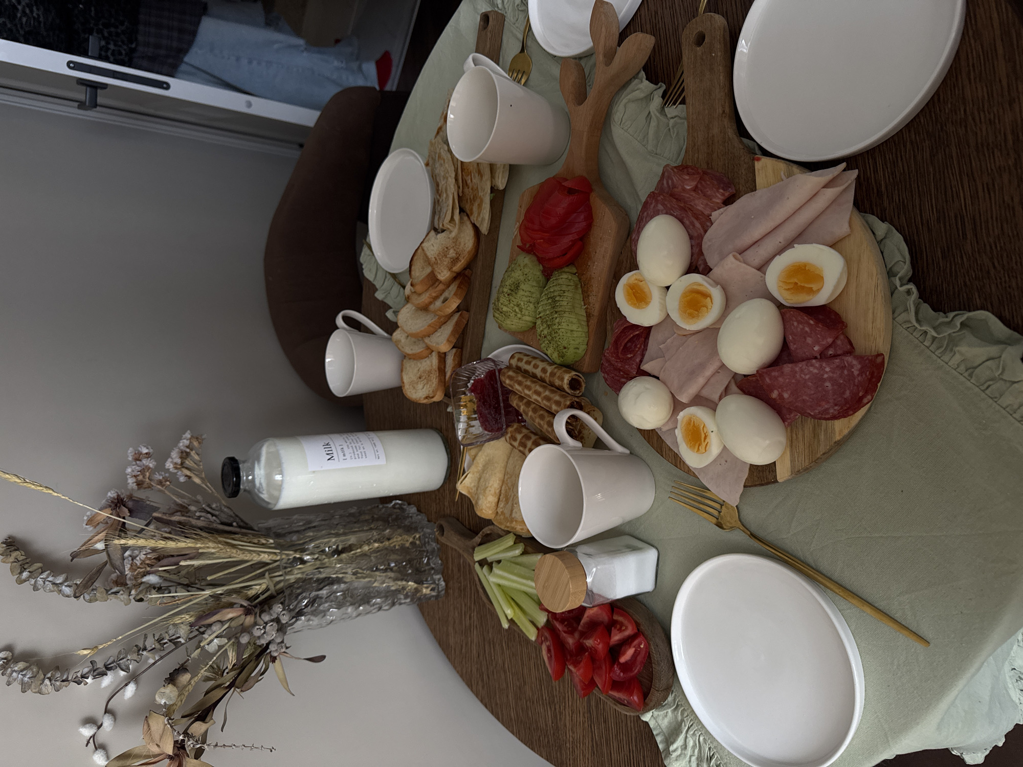 A high-angle shot displays a brunch arrangement on a dark wooden table covered with a light green tablecloth. The brunch includes two wooden boards: one holding sliced avocado, sliced red peppers, bread, and two white mugs, and the other holding hard-boiled eggs, ham, and salami. Accompanying the boards are additional plates, cutlery, a bottle of milk, a small container of salt, and a vase of dried flowers. The setting suggests an indoor dining area. There is no evidence to suggest that this photo was taken in Razvilka, Russia.