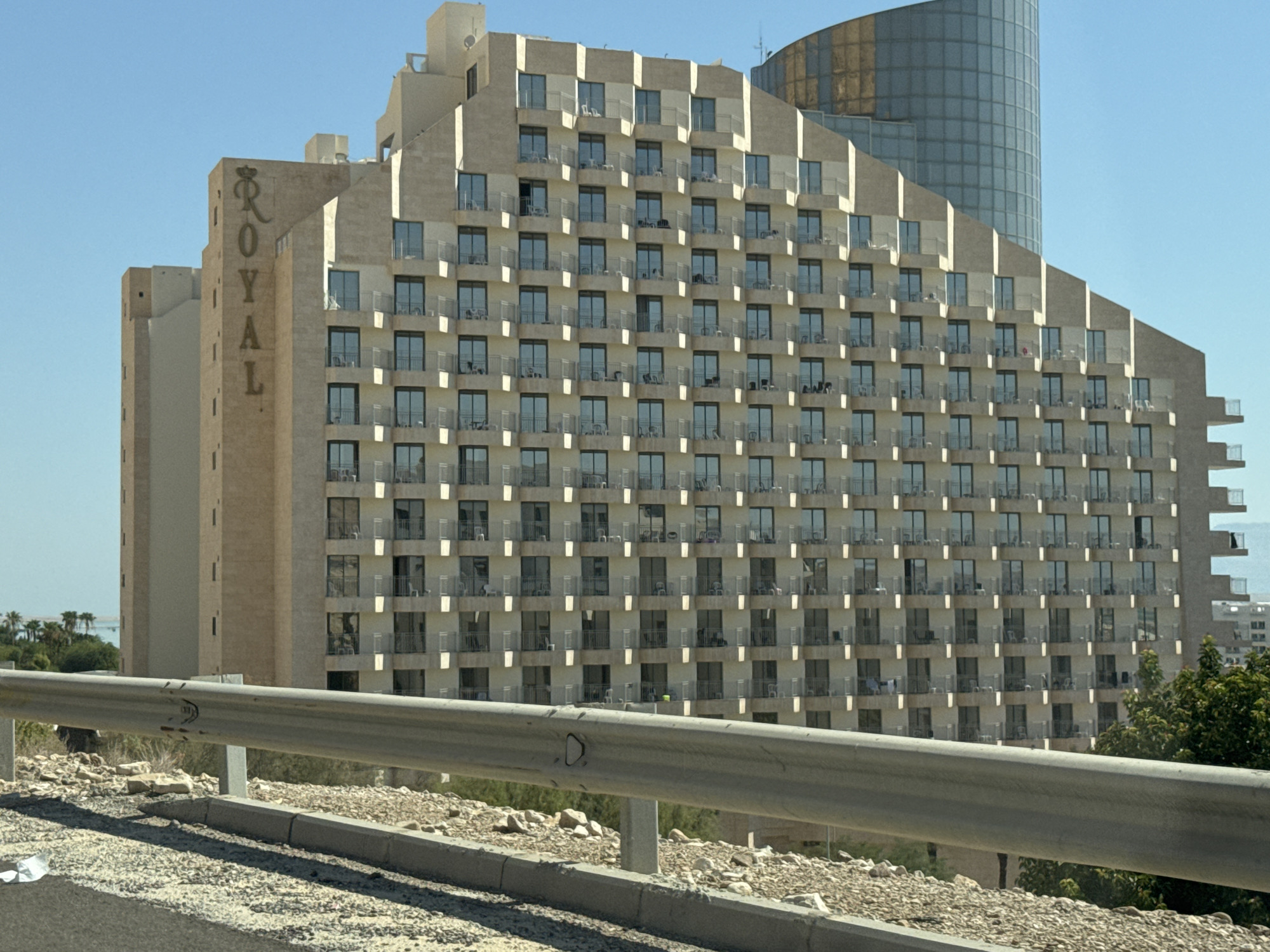 The image shows a large, modern hotel building in Tamar Regional Council, Israel, likely the "Royal" hotel, as indicated by signage on the left side of the building. The hotel is multi-storied with a repetitive facade of balconies and windows. To the right of the main hotel structure, a taller, cylindrical building with a glass facade rises above it. The hotel is situated in an arid or semi-arid environment, with rocky terrain and sparse vegetation visible in the foreground and background. A highway barrier runs across the bottom of the image, obscuring the immediate foreground. The sky is clear and blue. There are no visible people in the scene.