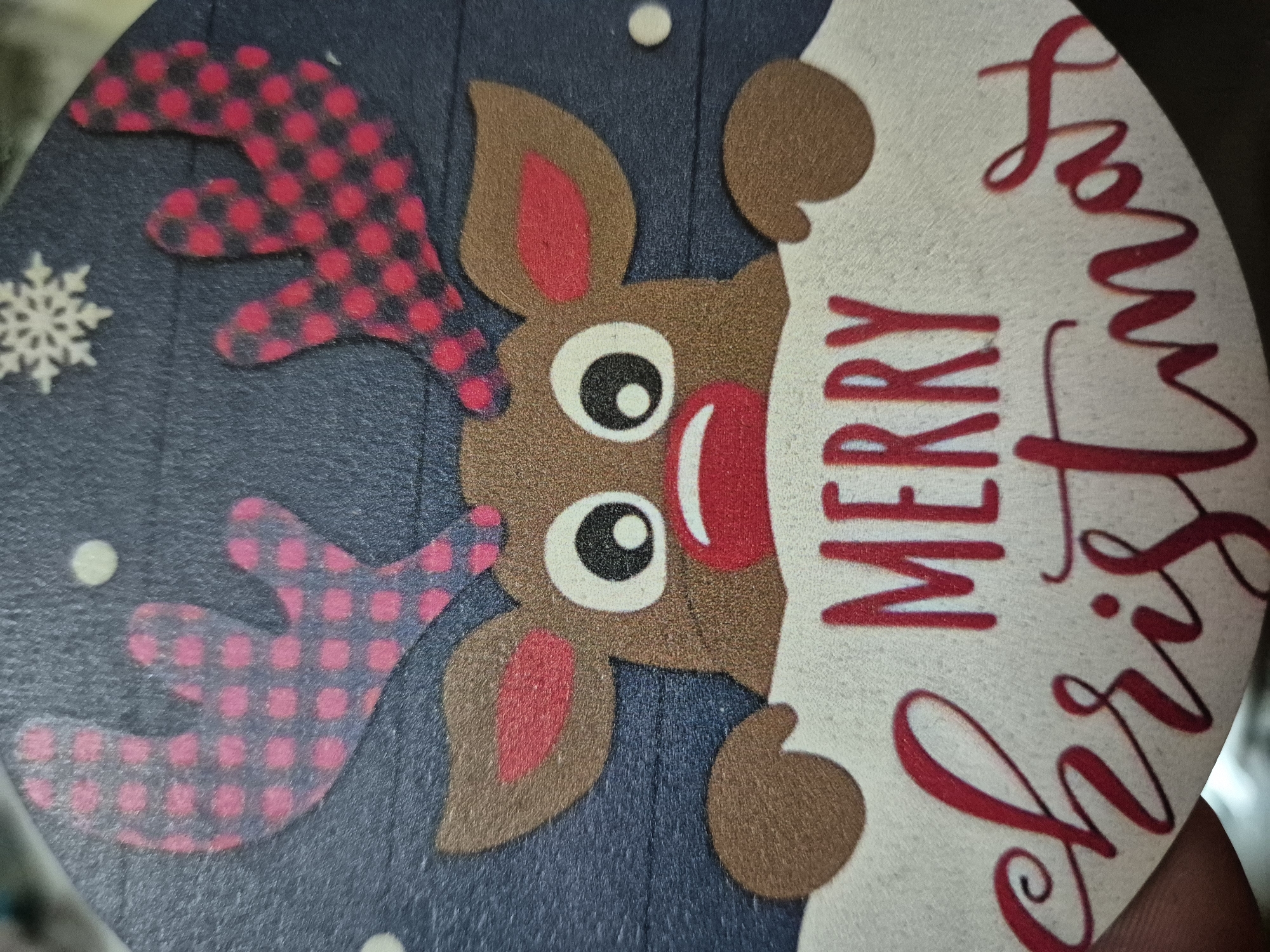 This image shows a close-up of a Christmas-themed object. The dominant feature is a cartoon-like reindeer with patterned antlers. The reindeer is brown, with red accents on its ears and nose, and big white eyes. The antlers have a red and black plaid pattern. The reindeer seems to be peeking over the edge of a white surface that has the words "Merry Christmas" written in red script. The background behind the reindeer is a dark blue, sprinkled with white dots and a snowflake.