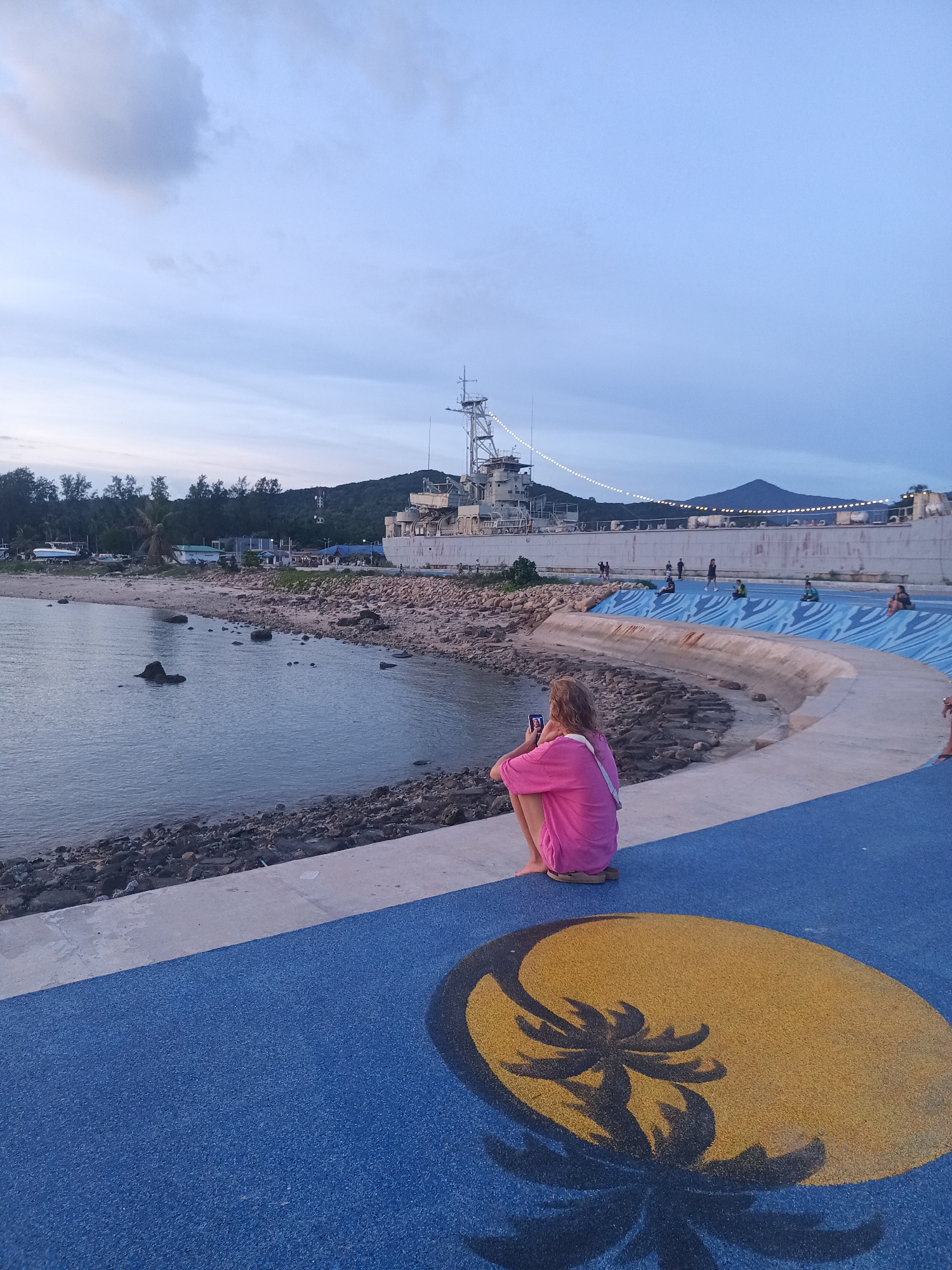 The image depicts a scenic view of a coastline in Koh Phangan, Thailand, during what appears to be dusk. The sky is a soft blend of blues and greys, with hints of clouds suggesting fair weather. In the foreground, a person is seated on a vibrant blue pathway, which is decorated with a circular motif of a palm tree silhouette against a yellow background. The person is wearing a bright pink outfit and is looking at their phone, possibly taking pictures or checking messages.
Beyond the pathway, a rocky shore meets the calm water of the sea, which reflects the muted tones of the sky. Scattered rocks are visible along the water's edge. Further back, a large, imposing ship, likely a museum or a permanent exhibit, dominates the background. Its structure suggests it's a historical vessel, possibly a warship, with various decks and a prominent mast. Lights are strung along the ship's railing, adding a festive touch to the scene, especially as daylight fades.
Several other individuals can be seen scattered along the pathway and the ship's vicinity, suggesting this is a public area for leisure and sightseeing. The overall atmosphere is tranquil, with a sense of calm leisure as people enjoy the coastal scenery and the presence of the ship. The location cues, such as the palm tree artwork and the tropical coastline, firmly place the scene in Thailand, specifically Koh Phangan. No visible text is present in the image.