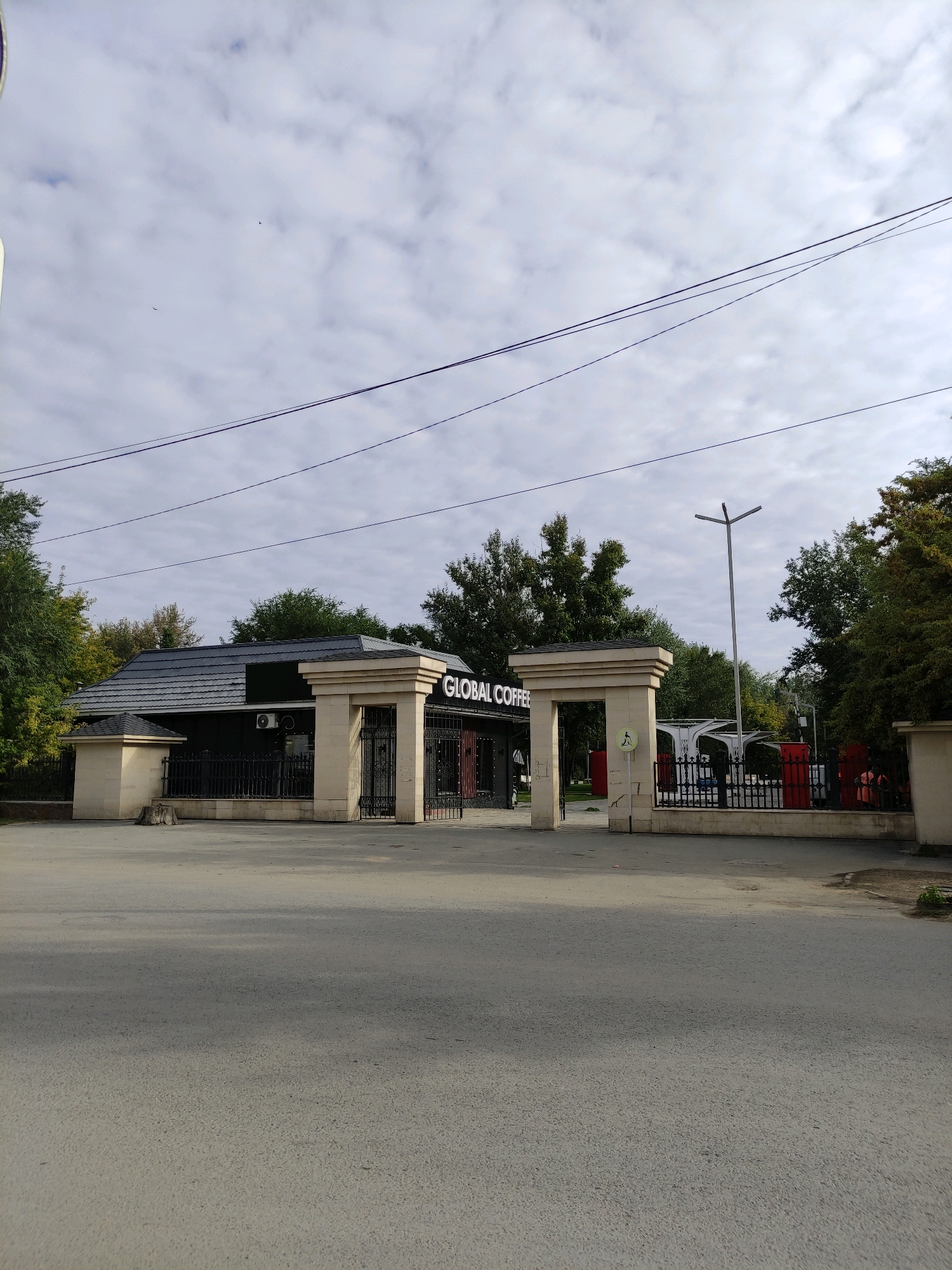 The image shows the entrance to "GLOBAL COFFEE" in Semey, Kazakhstan. The entrance features two large stone pillars framing an ornate black metal gate. Behind the gate, a dark-colored building with a grey, tiled roof is visible. The building has a sign that reads "GLOBAL COFFEE" in illuminated white letters. Trees with green and yellow foliage surround the building and entrance, suggesting it might be autumn. In the foreground, there is an asphalt road. The sky is overcast with some clouds. A modern-looking streetlight stands tall to the right of the entrance. A few people, appearing to be customers, are visible near the entrance, some entering and exiting the establishment.