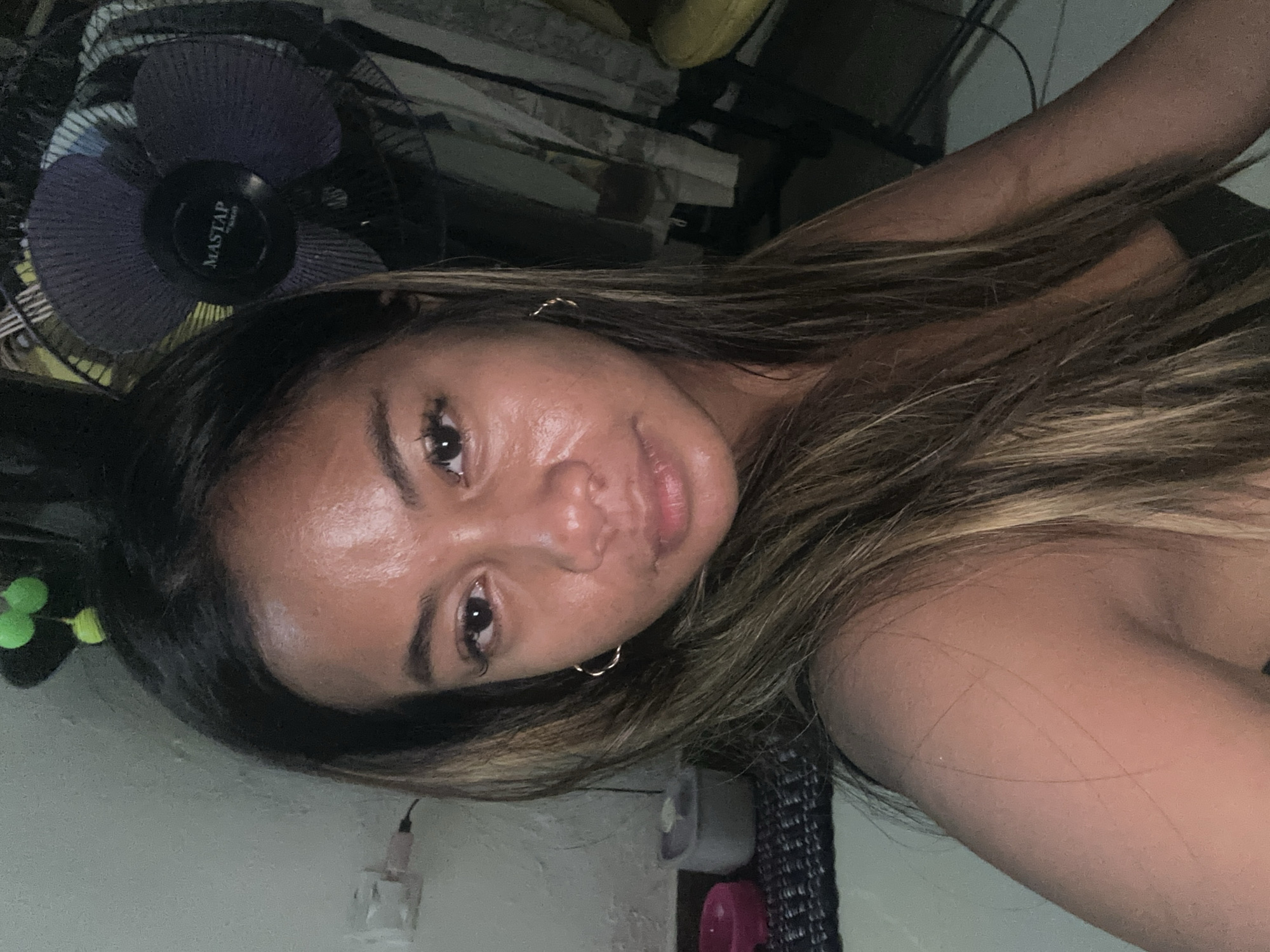 A young woman with long, brown hair and a light tan is looking at the camera. She is wearing a black top and gold hoop earrings. Her skin appears shiny, possibly from sweat or oil. In the background, a purple fan with the brand name "MASTAP" is visible, along with some household items. The lighting is dim, creating shadows. The overall setting appears to be indoors, potentially a bedroom or living area in Sukawati, Indonesia.