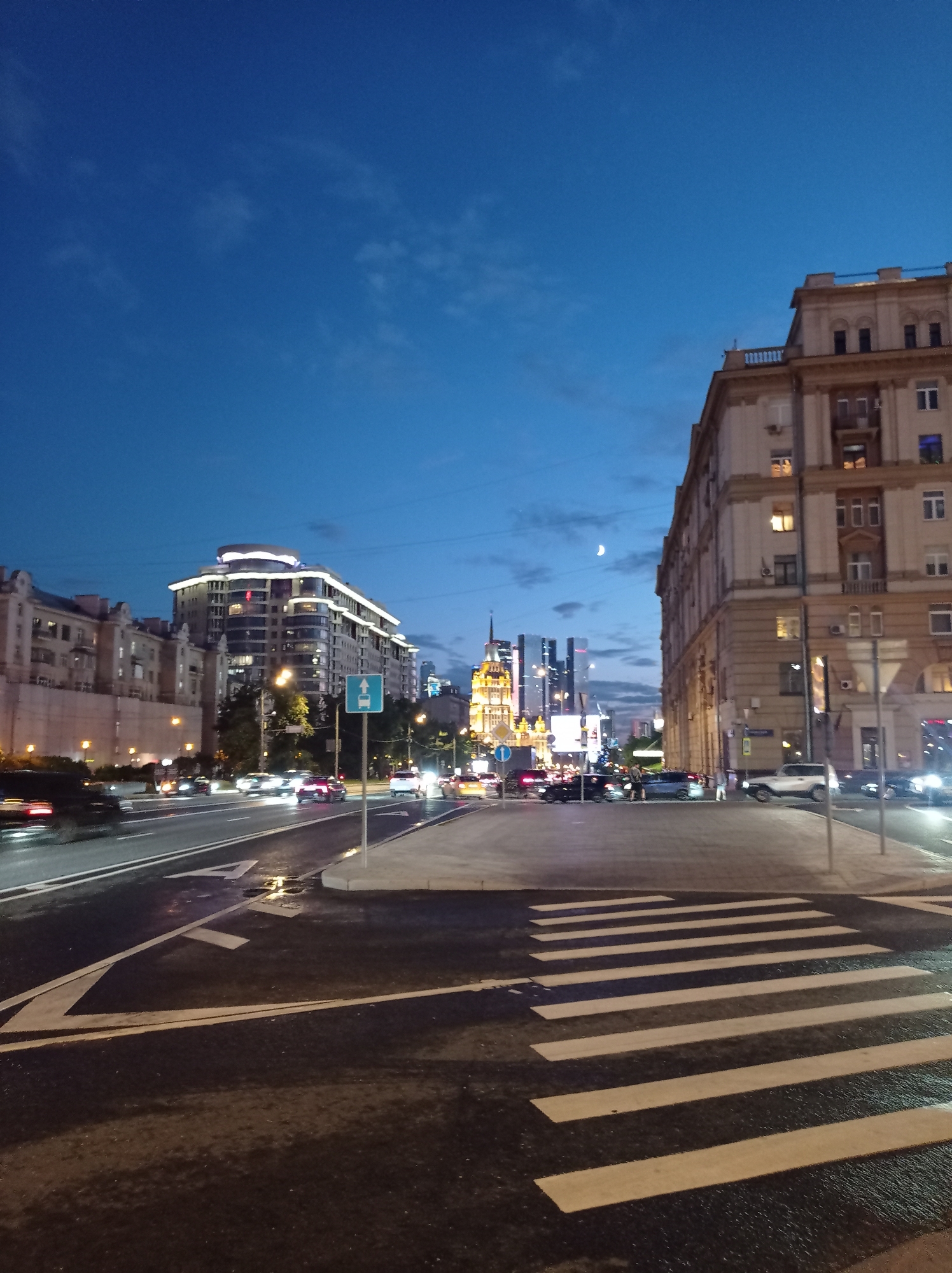 The image captures a wide-angle view of a city street in Moscow, Russia, at dusk. The sky is a deep blue with scattered, wispy clouds and a visible crescent moon. On the right, a large, ornate, beige building with multiple windows, some illuminated, dominates the foreground. To the left, a modern, curved building with extensive lighting stands out. In the mid-ground, several vehicles are in motion on a multi-lane road, their headlights and taillights creating light streaks due to the long exposure. A pedestrian crosswalk is clearly visible in the foreground, marked with white stripes on the dark asphalt. Several traffic signs are present, including one indicating a bicycle path. The streetlights are illuminated, casting a warm glow on the wet pavement, suggesting recent rain. In the distance, a cluster of taller, modern skyscrapers with illuminated windows are visible against the twilight sky. Two pedestrians are standing on the sidewalk near the crosswalk, interacting with each other. The overall scene depicts a bustling urban environment during the transition from day to night.