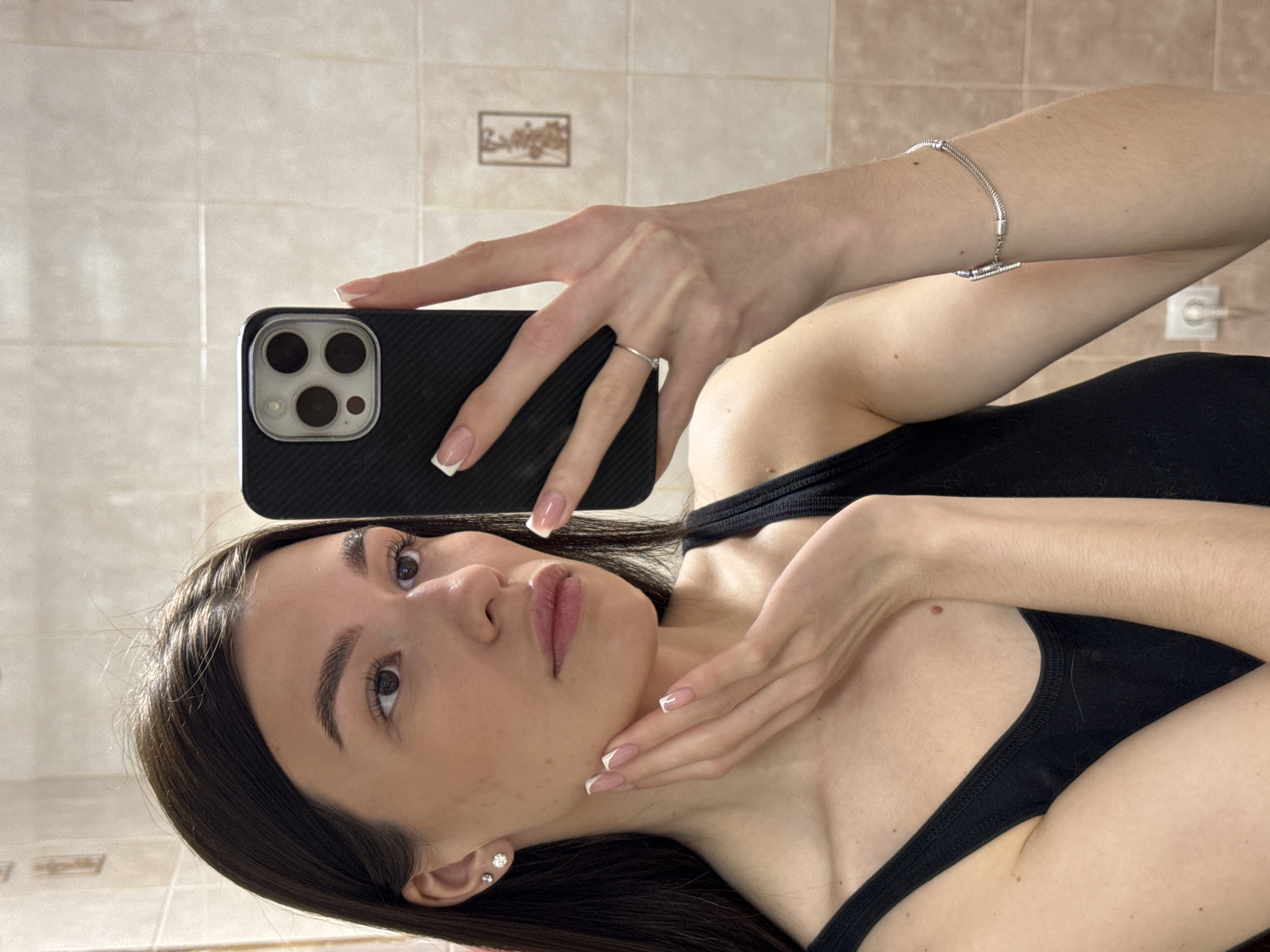 This media file is a mirror selfie, rotated 90 degrees clockwise, featuring a young woman in an indoor setting in Chaikovsky, Russia. The subject is a woman with long dark hair, dark eyebrows, and dark eyes, appearing to be in her late teens or early twenties. She wears a black tank top and has a composed expression, looking directly at the camera.
She is holding a dark-colored smartphone, possibly an iPhone, with a textured case, in her right hand. Her left hand is gently placed on her jawline and neck. She is adorned with a simple silver-toned ring on her left ring finger and a silver-toned chain bracelet with a small, sparkly charm on her right wrist. Her left ear features two small stud earrings. Her fingernails are manicured with a classic French tip design.
The background consists of light-colored, rectangular wall tiles, suggesting a domestic interior, possibly a bathroom. A small, framed decorative item, possibly a picture or relief with a tree motif, is visible on the tiled wall. An electrical outlet is also discernible in the background. No discernible text is visible in the image. The lighting is even, suggesting an indoor environment.