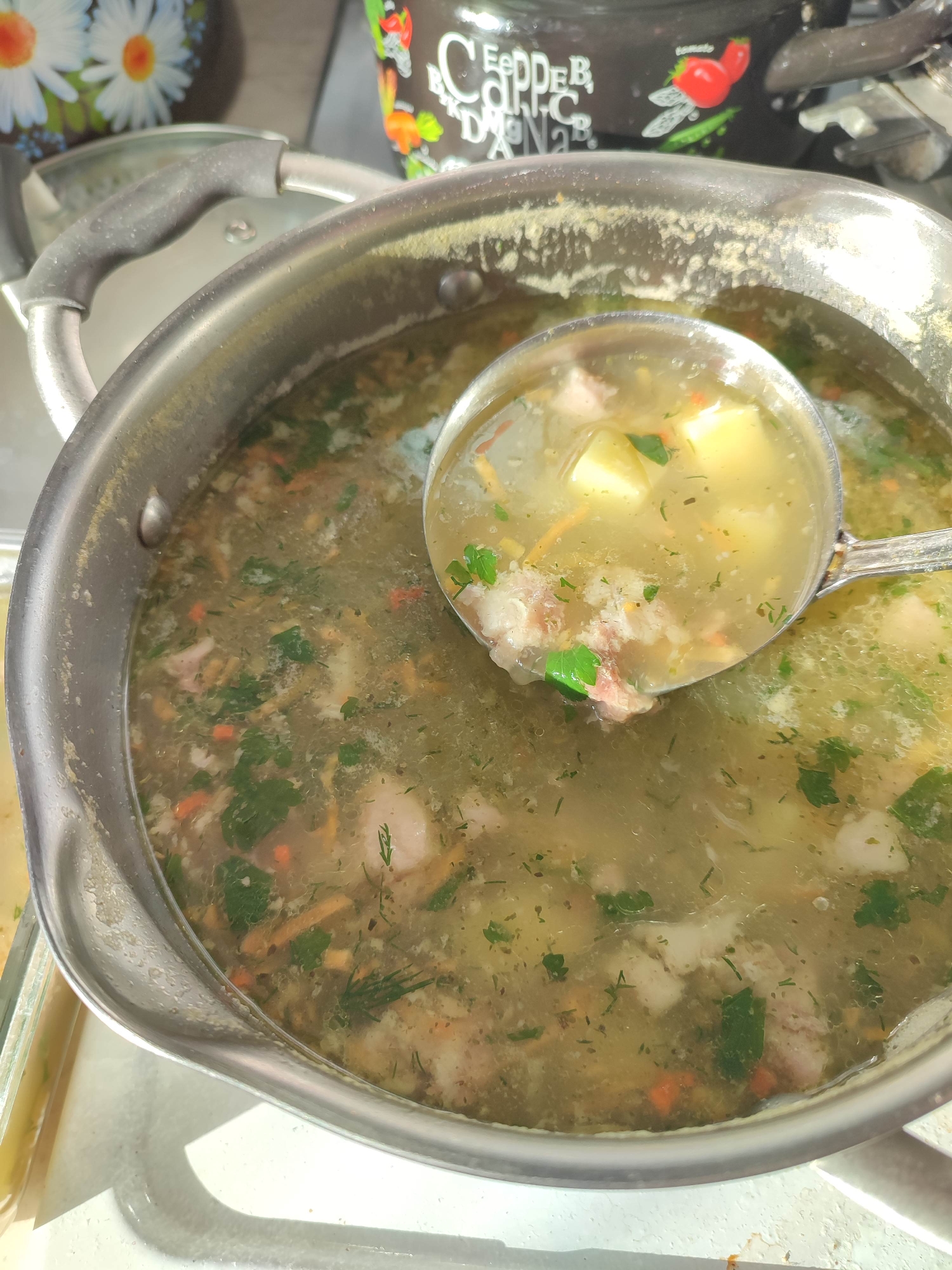 A top-down, medium-shot captures a pot of soup being served with a ladle. The soup appears to contain potatoes, meat, carrots, and green herbs, suggesting it is a broth-based soup. The stainless-steel pot has a handle, and a ladle scoops up some of the soup. Other containers are visible on the left side of the frame. The setting seems to be indoors, possibly a dining area or kitchen in Unknown, Unknown.