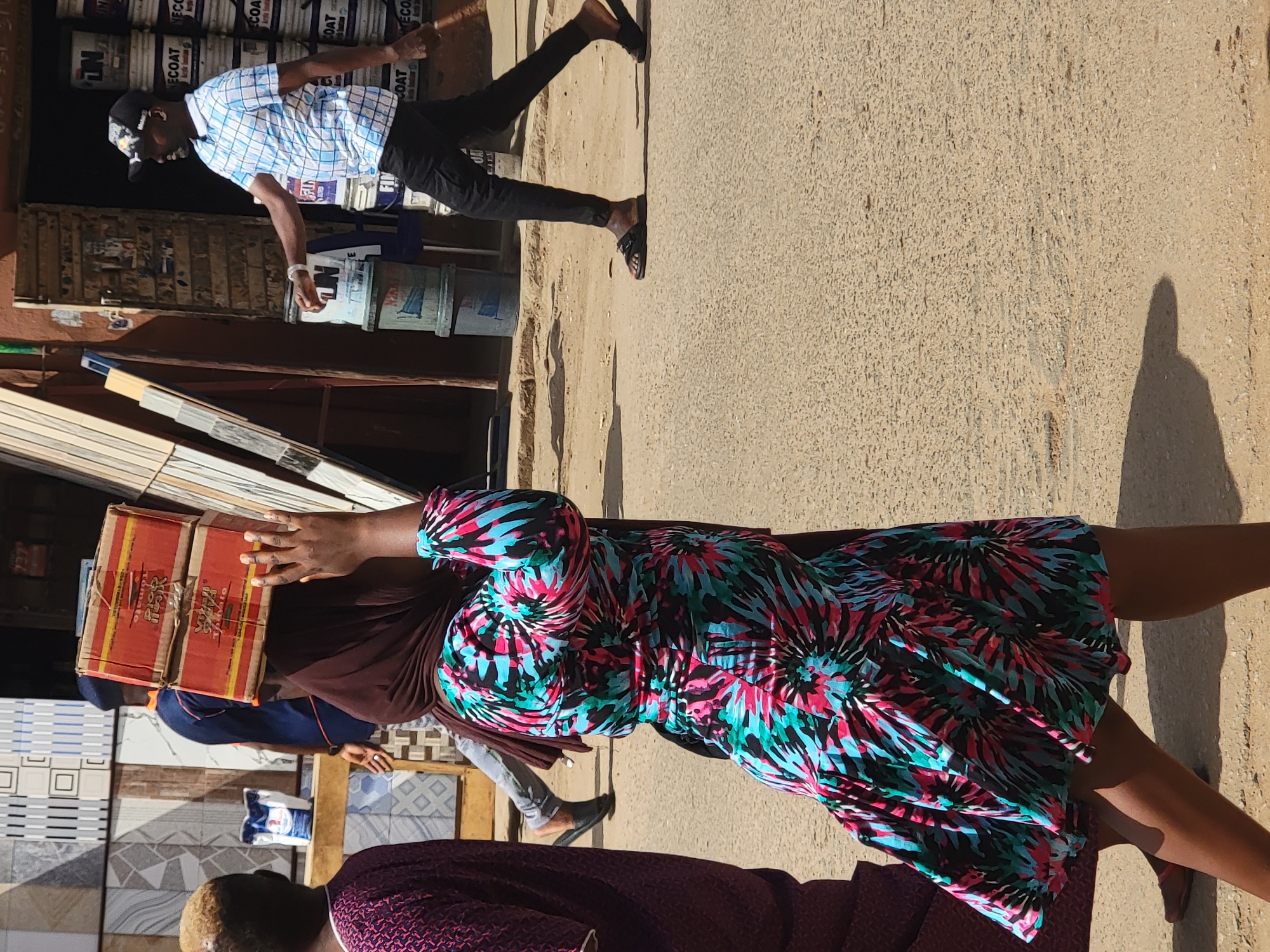The image shows a street scene in Maiburiji, Nigeria. In the foreground, a woman wearing a colorful dress is carrying a box on her head, obscuring her face. Behind her, part of another person wearing a patterned garment is visible. Next to her, a man is carrying tiles. The background features a shop selling paint and tiles, with a man in a checkered shirt walking in front of it. The scene is bathed in sunlight, casting shadows on the street.