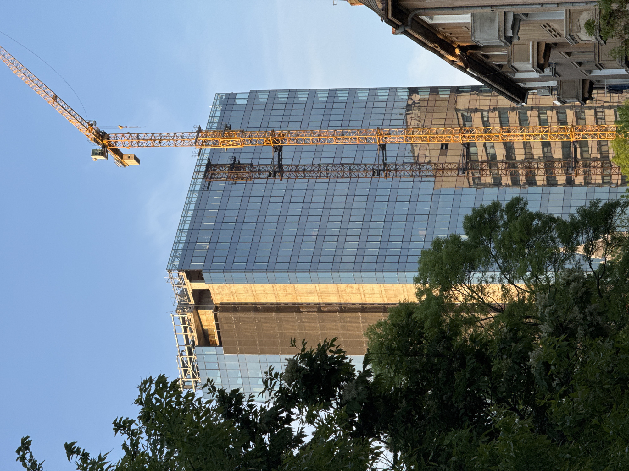 A low-angle shot captures a modern glass building under construction in Sofia, Bulgaria, seen against a clear blue sky. A yellow construction crane is prominently positioned on the left side of the frame, extending towards the top. The building's facade reflects the surrounding environment. Mature trees are in the foreground, partially obscuring the lower portion of the structure. The upper right corner shows a corner of an older, more traditional building. The scene is likely photographed during daylight hours.