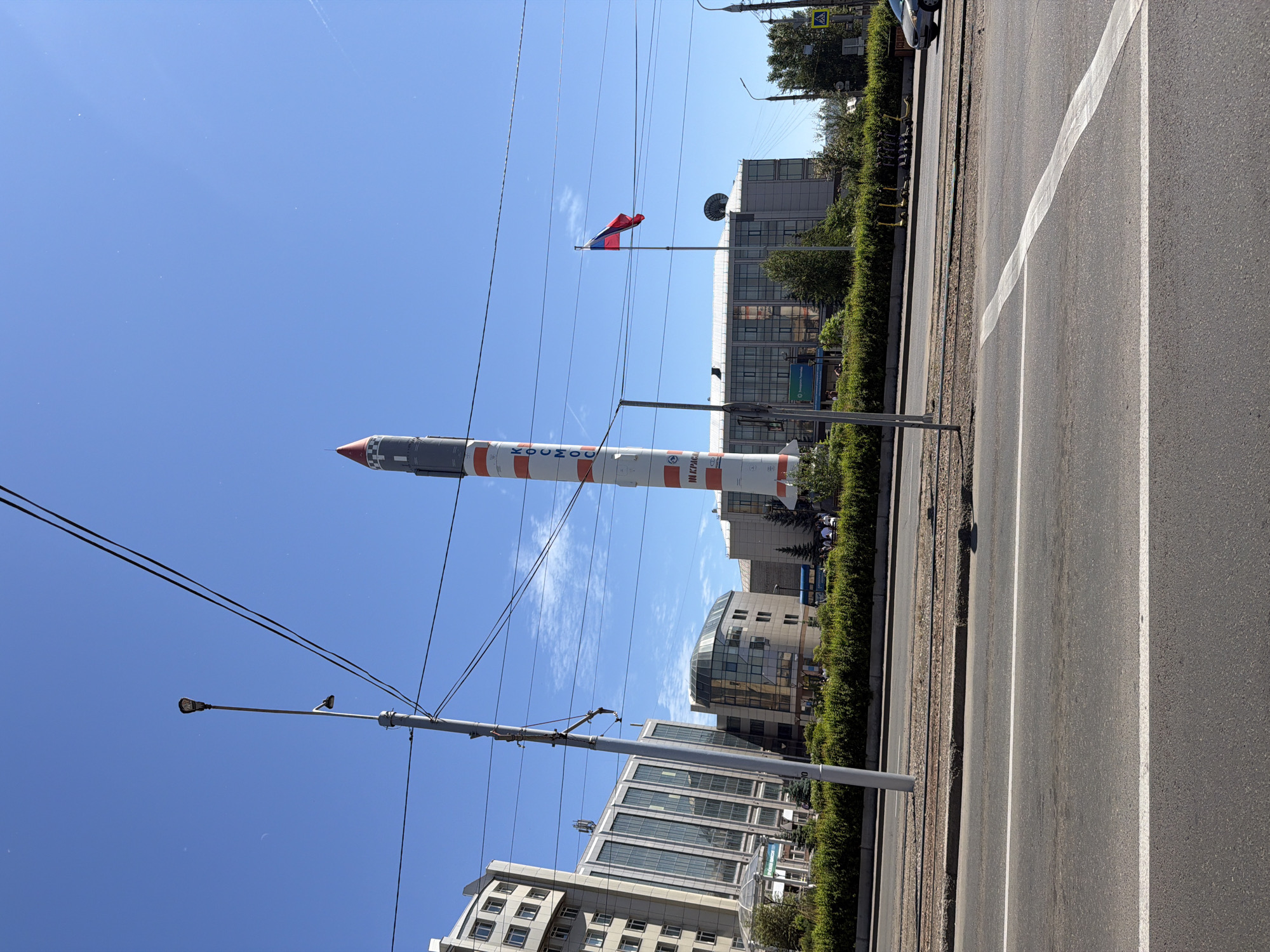 In Krasnoyarsk, Russia, a tall, monument-like rocket stands vertically on the left side of a street. The rocket is white with red and grey accents and has the Cyrillic letters "КОСМОС" written vertically on its side. To the right of the rocket, a Russian flag flies from a flagpole in front of a modern, multi-story building. Adjacent to this building are other buildings with glass facades and a green hedge runs along the street. A streetlamp is visible in the foreground, and overhead, thick electrical wires cross the frame. The sky is clear blue with a few wispy clouds. The street in the foreground shows clear lane markings. A few people are visible walking near the buildings and a line of scooters are parked on the sidewalk. A silver car is partially visible on the right side of the road. The overall scene depicts an urban environment on a bright, sunny day.