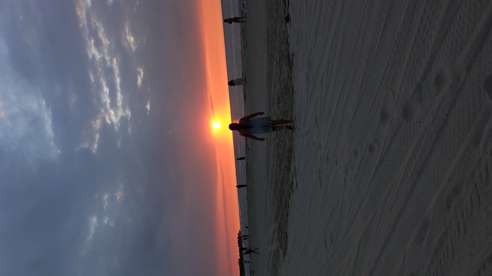 The image shows a sunset over a beach. A person stands on the sand, silhouetted against the setting sun. Other people are visible further down the beach. The sky is a gradient of colors, from dark blue and grey on the left to orange and red near the horizon. Tire tracks are visible in the sand. The location is likely Bocapan, Peru.