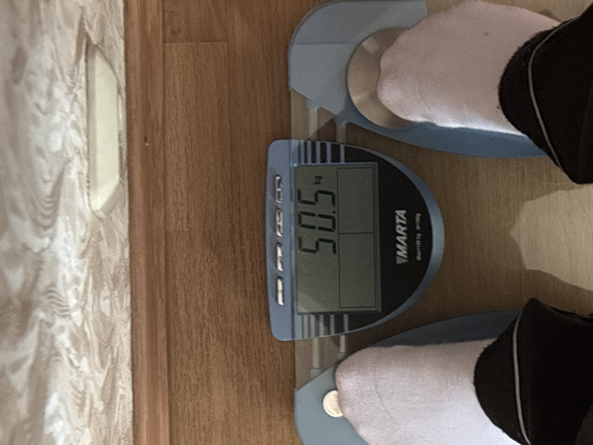 The image shows a close-up view of a person standing on a digital scale. The scale displays a weight of 50.5 kg. The person is wearing white socks and dark pants. The scale is blue and gray and has the brand name "MARTA" visible. The background consists of a wooden floor and a patterned wall. There is no indication of the city of Sinyavino, Russia, in this image.