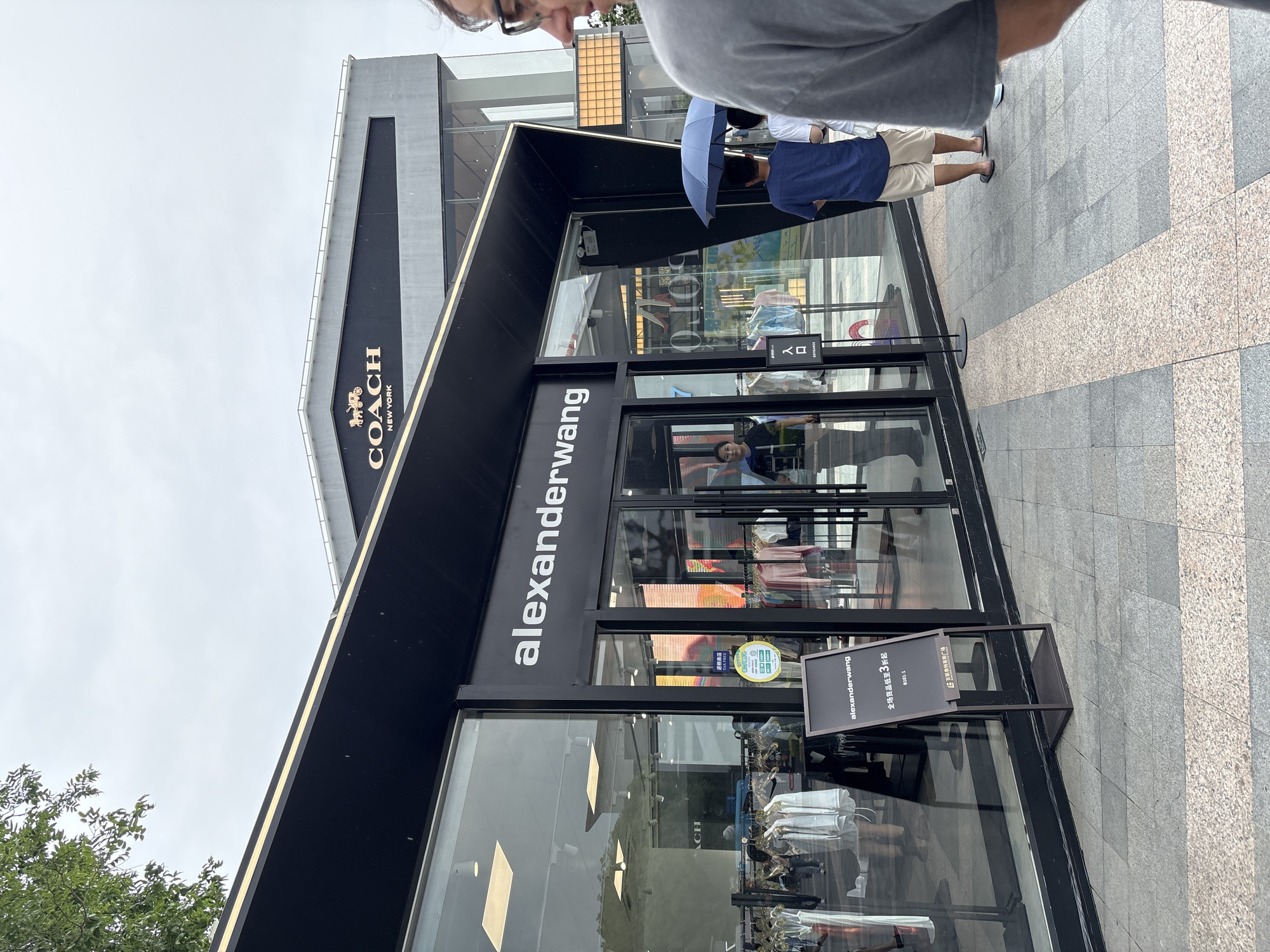 The image captures the exterior of a Coach store and an Alexander Wang store, likely situated within an outdoor shopping plaza in Qingpu District, China, on a cloudy day. The Coach store's prominent signage with its logo and "NEW YORK" text is visible on the upper facade of the building. Below it, the Alexander Wang store features a sleek, modern storefront with large glass windows showcasing merchandise and a prominent "alexanderwang" logo above the entrance. A black A-frame sign for Alexander Wang is placed on the sidewalk in front of the store, indicating a sale. Several individuals are seen walking and standing outside the stores, some holding umbrellas, suggesting it may be raining or overcast. The ground is paved with large square tiles. Reflections in the glass windows reveal the interior layout and displays of the stores, including clothing racks.