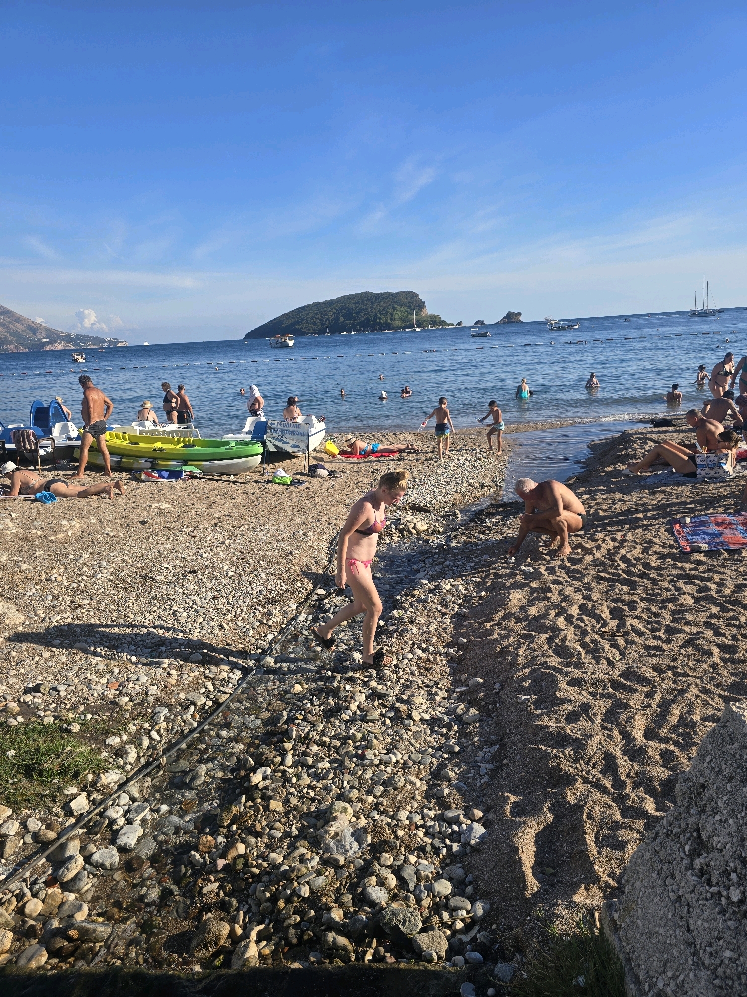 In Budva, Montenegro, a crowded pebble and sand beach stretches along the Adriatic Sea under a clear blue sky. The water is calm, with several boats dotting the horizon and a small island visible in the distance. On the beach, people are engaged in various activities. Some are sunbathing on towels and loungers, while others are wading in the shallows. A group of children is playing near the water's edge, and a few individuals are walking along the shoreline. Several pedal boats and kayaks are pulled up onto the beach, indicating recreational opportunities. A small stream of water flows from the land out to the sea across the beach. The scene is typical of a summer day at a popular tourist destination.