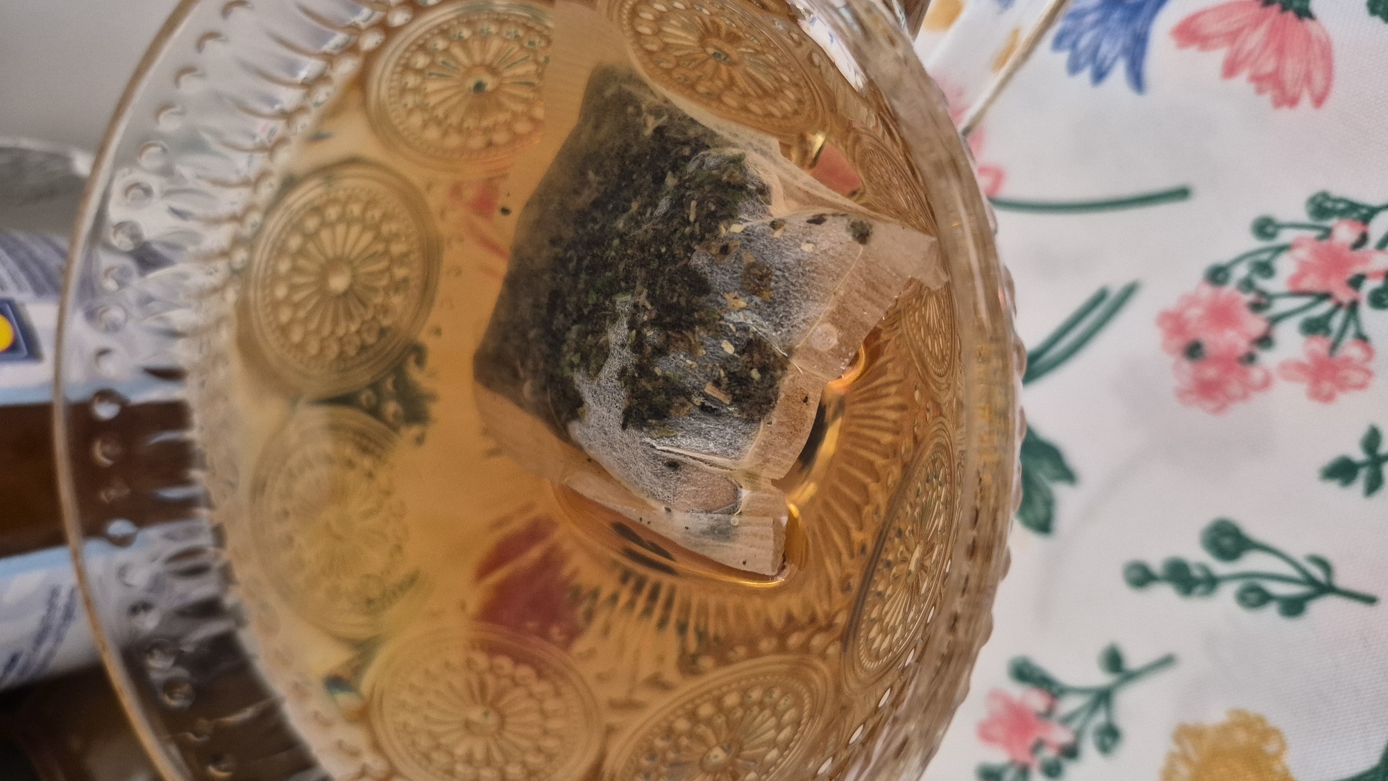 A close-up, overhead view of a glass teacup filled with amber-colored tea. A single, light-colored tea bag, containing dark green tea leaves and lighter flecks, is submerged in the tea, partially obscuring the decorative circular patterns embossed on the inside of the teacup. The teacup is placed on a white tablecloth adorned with a pattern of pink, yellow, and blue flowers and green stems. The background is blurred, but appears to show a light-colored bottle or container. There is no visible indication of the city of Krauja, Latvia.
