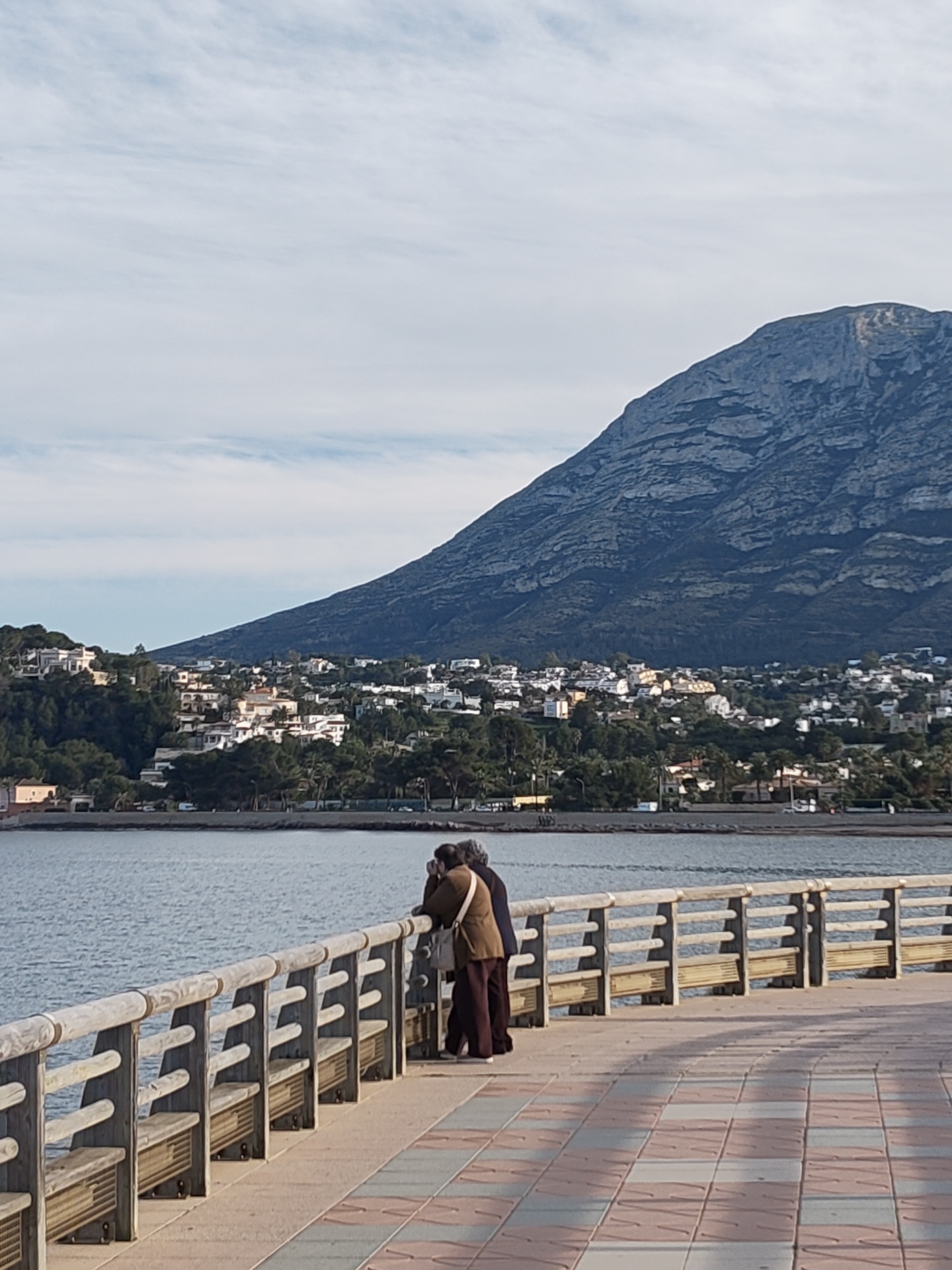 The image displays a coastal scene in Dénia, Spain, with the prominent Montgó Massif in the background. In the foreground, a paved promenade, featuring a pattern of alternating light and dark rectangular tiles, extends from the right edge, slightly curving to the left. A wooden railing system, incorporating benches, lines the promenade's seaward edge. Two individuals stand at this railing on the left, facing away from the viewer, looking out over the water. One person wears a brown jacket, dark trousers, and a light-colored shoulder bag. The second individual, with grey hair, wears a dark jacket and dark trousers, resting their arms on the railing. Beyond the promenade, the calm sea stretches to a distant coastline. A residential area with numerous white and light-colored buildings is situated along the lower slopes of the Montgó mountain, interspersed with green trees and vegetation. The large, rocky Montgó mountain occupies the background, its rugged terrain rising sharply under a sky with scattered white clouds.