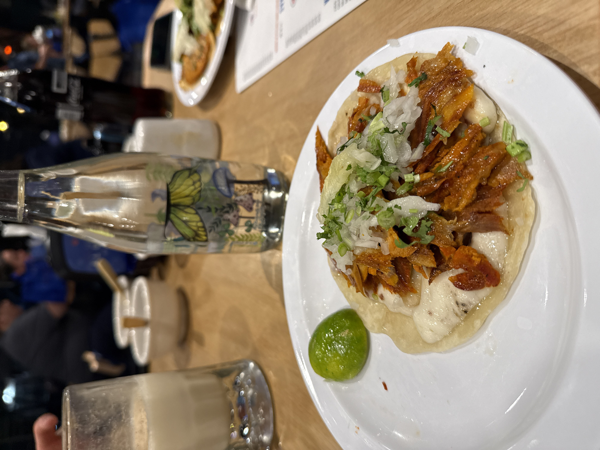The image shows a taco al pastor on a white plate, garnished with white onions, cilantro, and a lime wedge. The taco sits on a light-colored wooden table next to a glass bottle with a butterfly design, a glass with a beverage, and a small white cup with wooden sticks. In the blurred background, there are other plates, bottles, and people seated at the table. Based on the typical cuisine and presentation, the scene is likely in a restaurant or eatery in the city of Querétaro, Mexico.