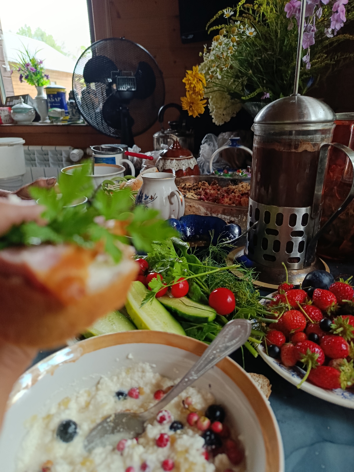 An overhead shot captures a rustic table laden with a fresh breakfast spread, set against a backdrop of a country home's interior. In the foreground, a slice of bread topped with salmon and greens is held up, its texture and freshness evident. To the right, a white bowl overflows with a cottage cheese-like dish, studded with blueberries and red currants, a spoon resting within. Adjacent to it, a platter overflows with vibrant strawberries, some blackcurrants, and plums, artfully arranged with fresh dill and parsley. Cherry tomatoes and cucumber slices also feature on the table. A French press filled with dark coffee stands tall, and in the background, a bouquet of mixed flowers, including daisies and yellow blossoms, adds a touch of natural beauty. A black oscillating fan and kitchenware are visible on a windowsill. The scene suggests a leisurely breakfast in a cozy, rural setting.