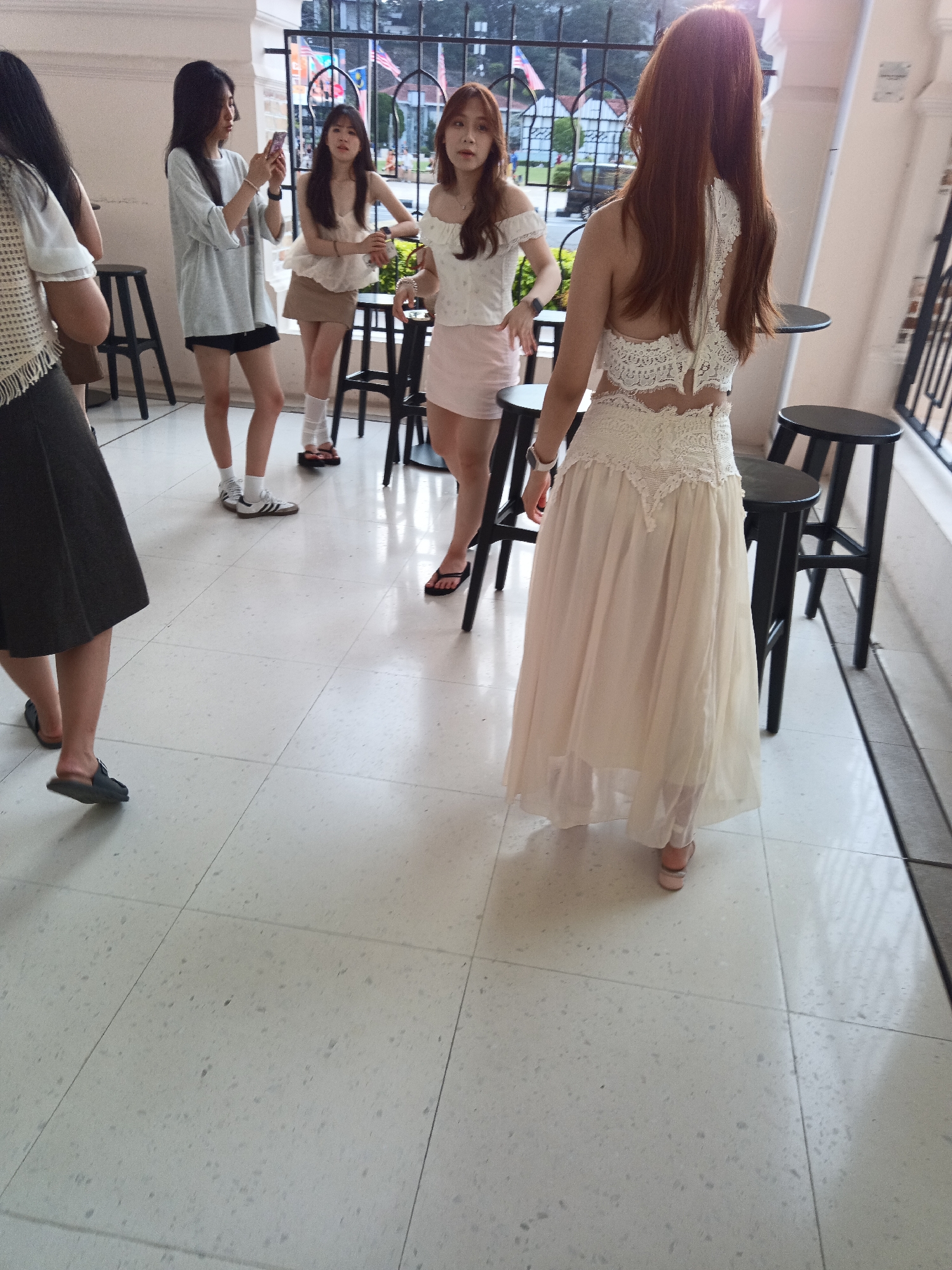 In Kuala Lumpur, Malaysia, four young women are gathered near an outdoor seating area, possibly a cafe or restaurant, with a barred fence in the background. The foreground is dominated by a cream-colored tiled floor with a speckled pattern. To the right, two black bar stools are visible, one with its legs extending towards the viewer.
The woman closest to the viewer is wearing a long, flowing, cream-colored dress with intricate white lace detailing on the back and bodice, and she is looking away from the camera. Next to her, another woman in a white off-the-shoulder top and a light pink skirt stands facing left, holding a drink. Further to the left, a woman in a grey t-shirt and black shorts is holding up a smartphone, seemingly taking a photo or video. She is wearing white socks and athletic shoes. Another woman, partially obscured by the first, wears a white top with a textured fringe and a dark skirt, and is facing to the right.
The environment appears to be an outdoor walkway or patio, with a white wall on the left and a building structure behind the fence. The fence itself is black and extends across the middle ground, with flags visible in the distance. The overall lighting suggests it is daytime, with soft light illuminating the scene.