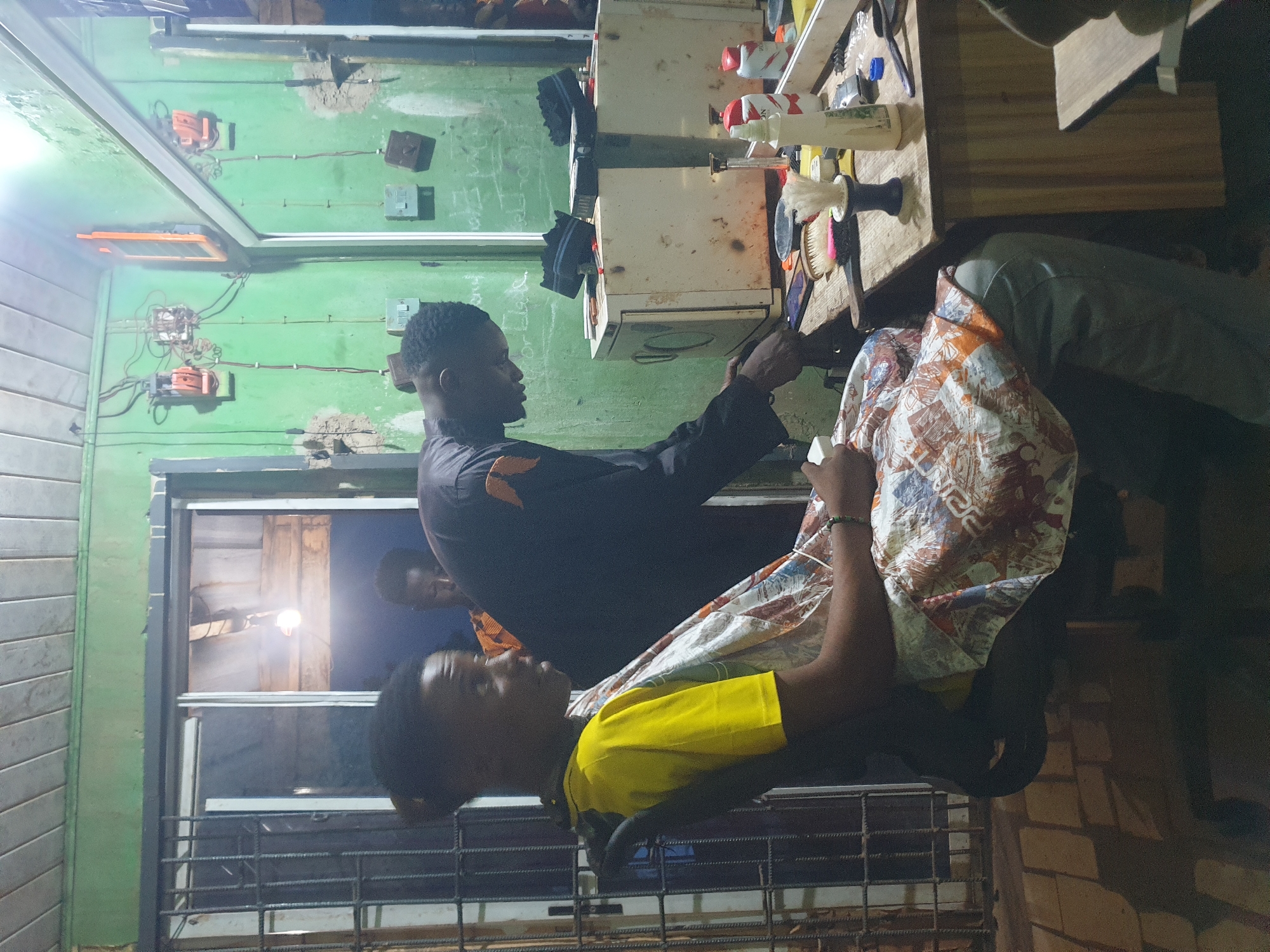 In Oluawo, Nigeria, a barber is cutting the hair of a young man seated in a chair. The barber, wearing a black shirt with an orange design, is actively engaged in his task. The customer, dressed in a yellow t-shirt and covered by a patterned cloth, is seated. Another individual, partially visible, stands behind the barber. The environment appears to be an open-air barbershop with rustic wooden elements, including a counter laden with barber tools and hygiene products. The walls are painted green, and electrical conduits are visible, suggesting a basic setup. The lighting is dim, with a single bulb providing illumination.