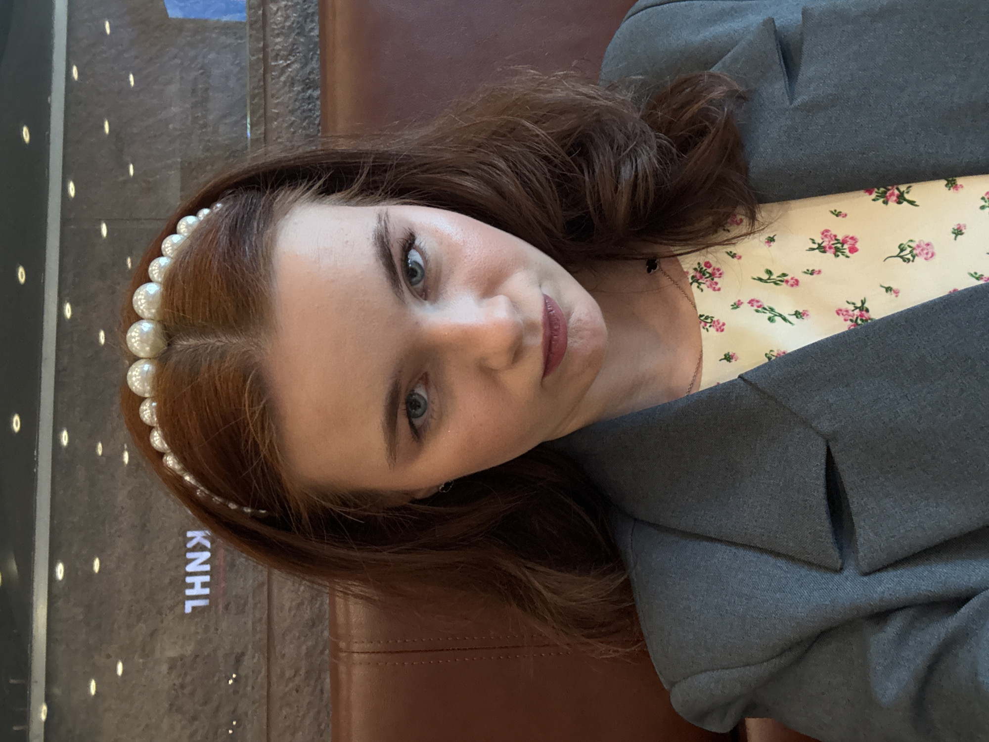 A young woman with reddish-brown, wavy hair, parted down the middle, is depicted in a medium close-up, rotated 90 degrees clockwise. She has blue-grey eyes and a subtle, soft smile on her lips, which are colored with a muted berry shade. She wears a white pearl-studded headband across the top of her head. Her skin shows a natural sheen.
She is dressed in a dark grey blazer or jacket over a light, possibly cream-colored, top adorned with a delicate pattern of small pink and green floral sprigs. A thin necklace with a small dark charm is visible around her neck.
The background consists of two main elements. To her right (bottom of the rotated image) is a rich, dark brown, upholstered surface, possibly leather, with visible stitching. To her left (top of the rotated image) is a dark, textured wall featuring a vertical array of small, glowing white oval or rectangular lights. On this dark wall, inverted due to the image orientation, the letters "KNHL" are visible in white text. The scene appears to be indoors under artificial lighting.