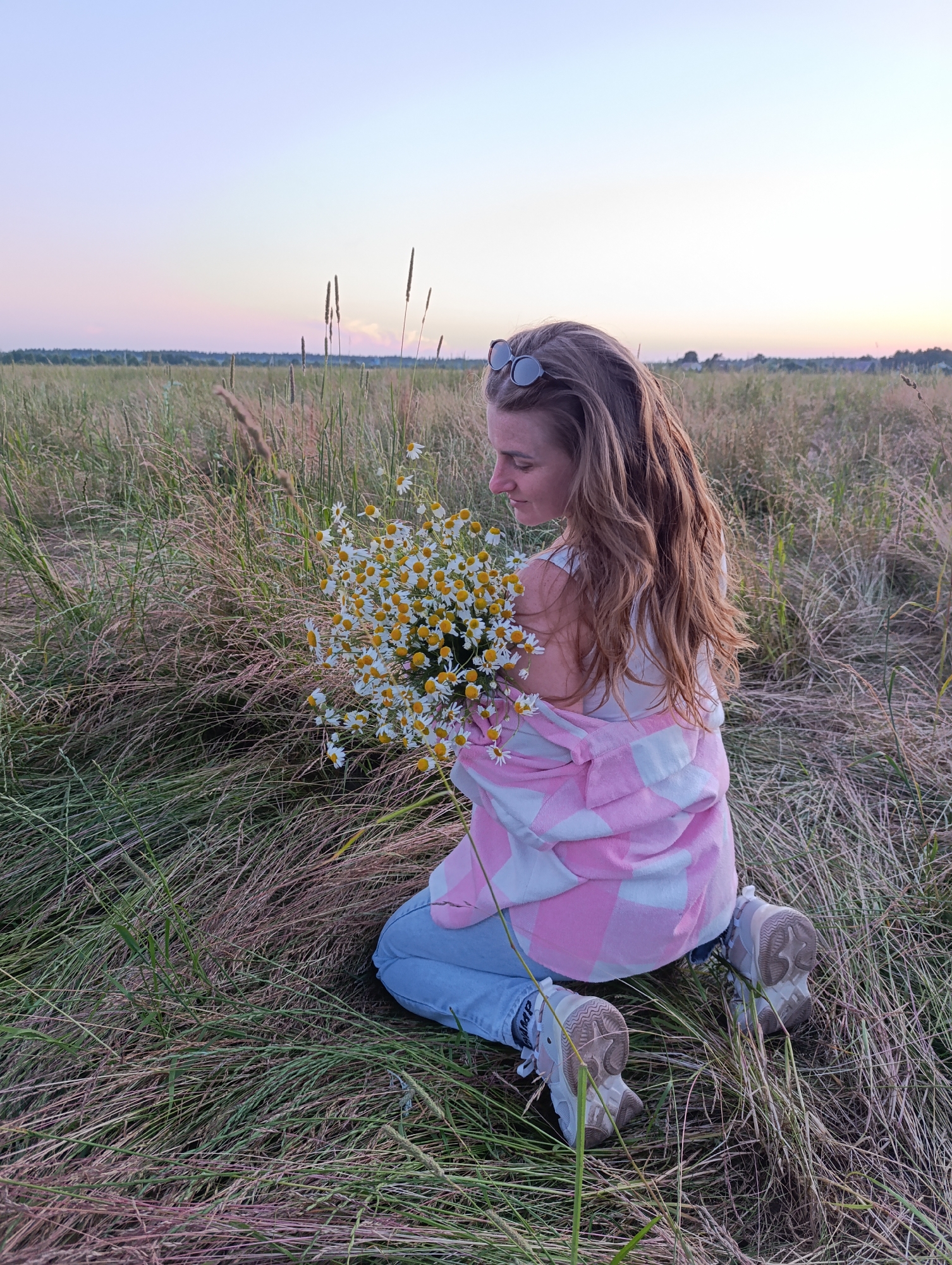 A woman with long, wavy brown hair, wearing sunglasses on her head, is kneeling in a grassy field. She is holding a bouquet of daisies with yellow centers. She is wearing a white tank top, blue jeans, and white sneakers with light brown accents. A pink and white checkered jacket is draped over her shoulders. The field is filled with tall, dry grass and some wildflowers. In the background, a hazy horizon with trees suggests a rural landscape. The sky is a soft gradient of pale blue, pink, and purple, indicating it is either sunrise or sunset. The lighting is soft and diffused, creating a serene and peaceful atmosphere. The woman appears to be looking down at the flowers with a contemplative expression.