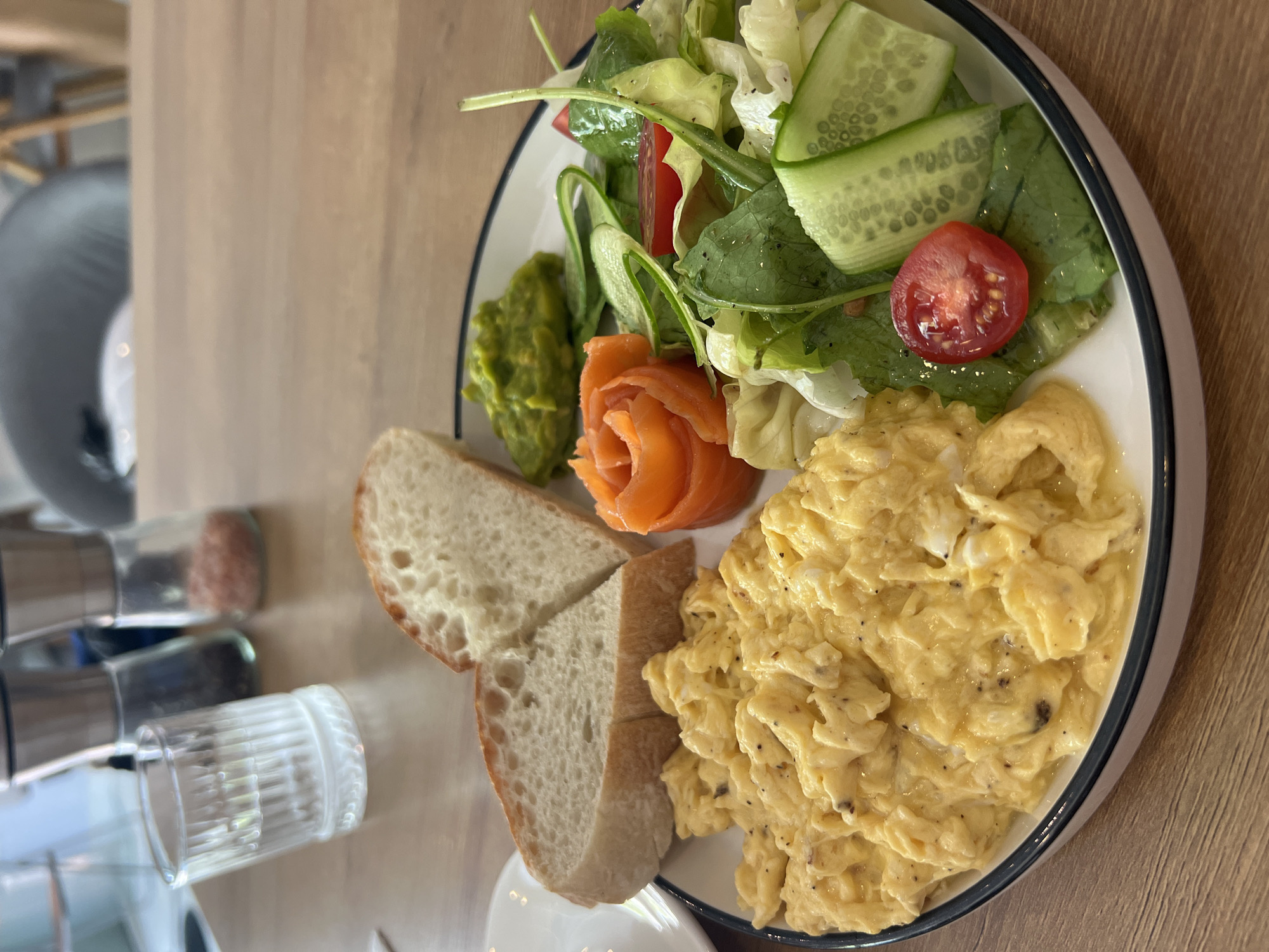 The image shows a plate of food on a wooden table. The plate contains scrambled eggs, two slices of bread, a salad with cucumbers and tomatoes, avocado, and smoked salmon formed into the shape of a rose. Salt and pepper shakers and a glass of water are visible on the table. The location is Bắc Nha Trang, Vietnam.