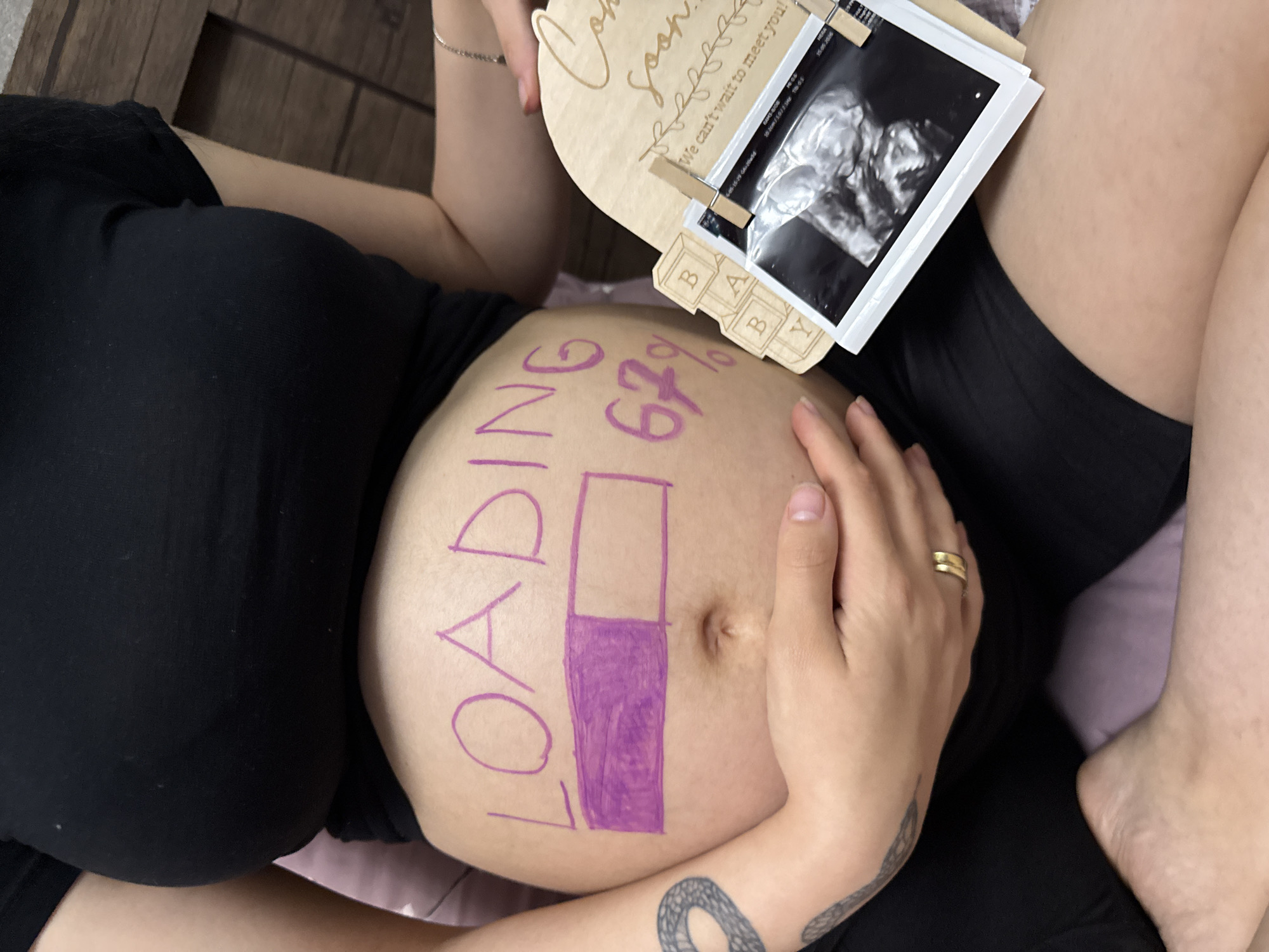In an indoor setting, likely a bedroom, a pregnant person is captured from the chest down. They are wearing a black t-shirt and black shorts. The person's pregnant belly is prominently displayed and decorated with purple marker. The word "LOADING" is written across the top of the belly, and a progress bar is drawn below it, filled approximately 67% of the way. To the right of the belly, a wooden decorative item with the words "Coming Soon" and "We can't wait to meet you!" is visible. Attached to this wooden item are several ultrasound photos clipped with small clothespins. The person's hands are resting on their belly, with a gold ring visible on one finger. The background consists of a wooden headboard and a soft pink surface. No specific location in Tbilisi, Georgia, can be identified from the image.