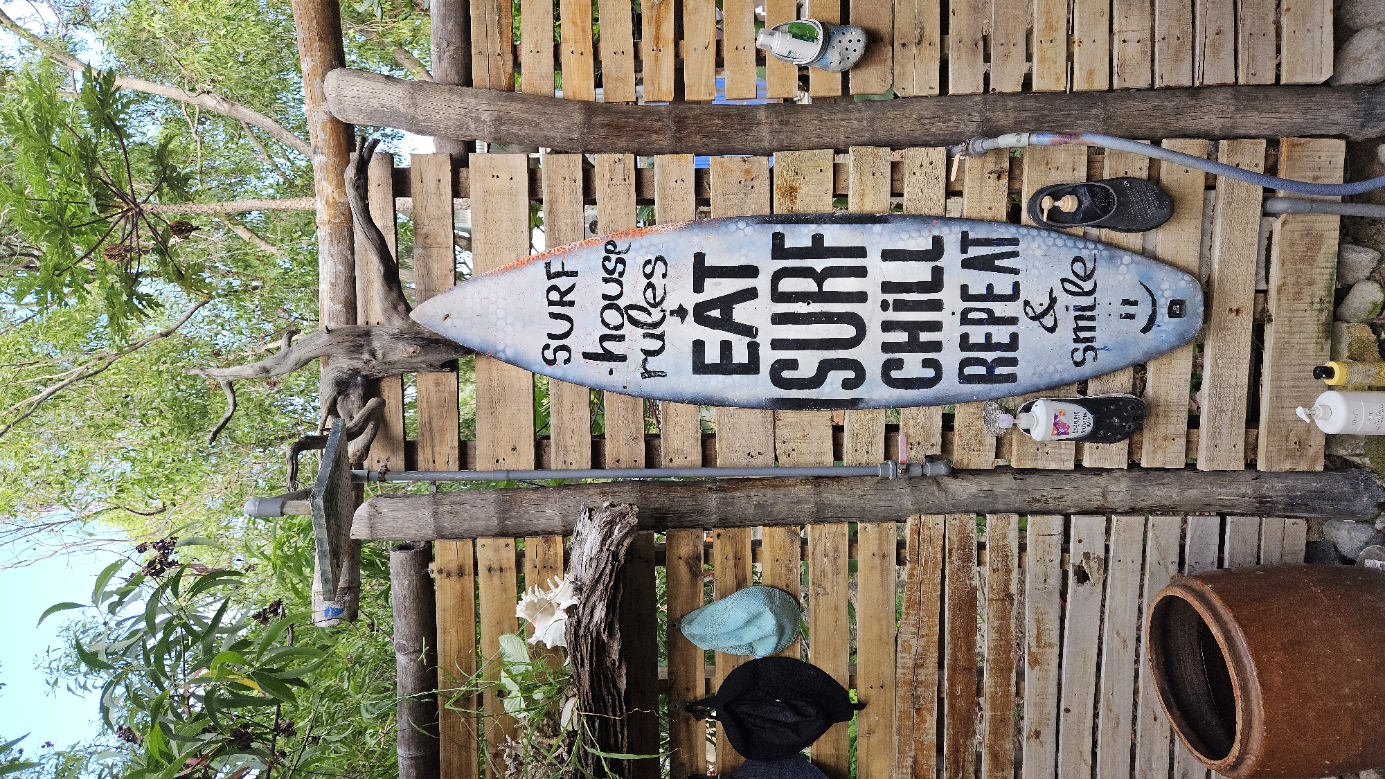 The scene presents a vertically oriented shot, capturing an outdoor shower area with rustic construction. The location is specified as Cam Lam, Vietnam. A surfboard with the text "SURF - HOUSE RULES EAT SURF CHILL REPEAT & SMILE :)" is mounted horizontally on a wall made of wooden pallets. The wall also holds an assortment of objects, including a pair of Crocs, a pair of flip-flops, bottles of toiletries, and several hats. A gray water hose is present along the top. A metal showerhead is positioned on the left side of the frame, connected to plumbing. Natural elements are visible including tree branches and foliage surrounding the structure. A large clay pot rests in the lower right corner.