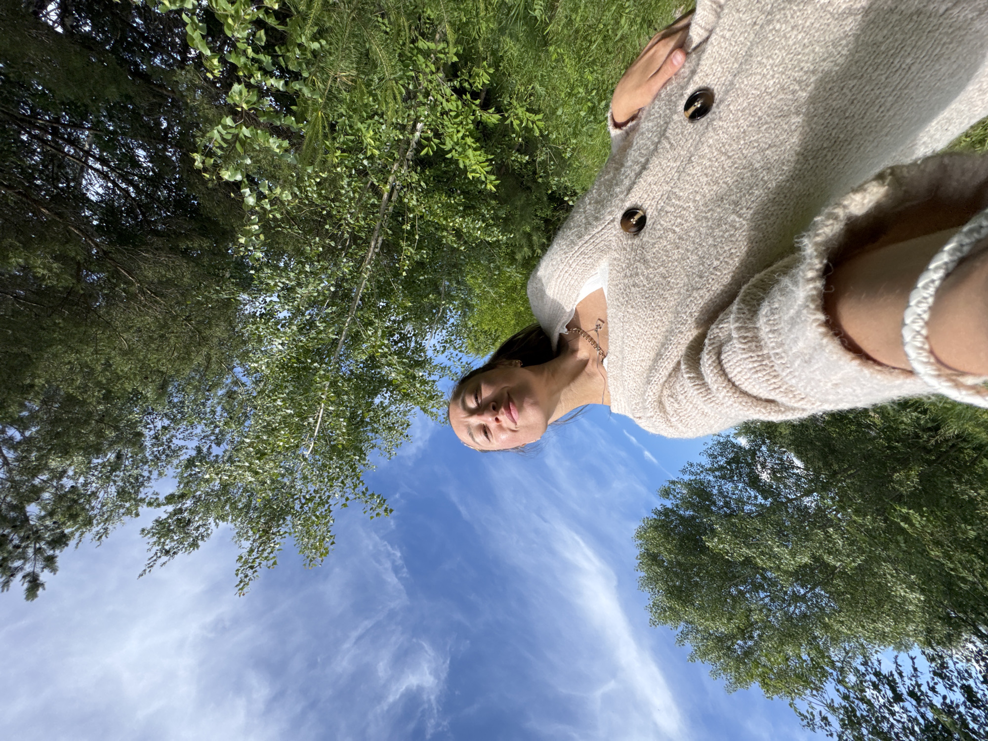The image displays a rotated, low-angle outdoor selfie of a woman in a natural setting. The image is rotated approximately 90 degrees clockwise. When viewed in an upright orientation, the woman's head is positioned in the lower-left quadrant, looking upwards towards the camera with a serene and relaxed expression, eyes slightly squinted and a subtle smile. She is wearing a light-colored (beige or cream) knit cardigan with two prominent brown buttons, a delicate necklace, and a light-colored braided bracelet on her left wrist.
The background primarily consists of a vast blue sky with scattered, wispy white clouds, indicating fair weather and bright daylight. Lush green foliage and tree canopies frame the upper and left portions of the scene, suggesting a wooded area or forest environment. Both coniferous and deciduous trees are visible. The low-angle perspective provides an immersive view looking up through the trees towards the sky. The activity appears to be enjoying the outdoors, possibly within a forest or park in Chaikovsky, Russia. There is no visible text.