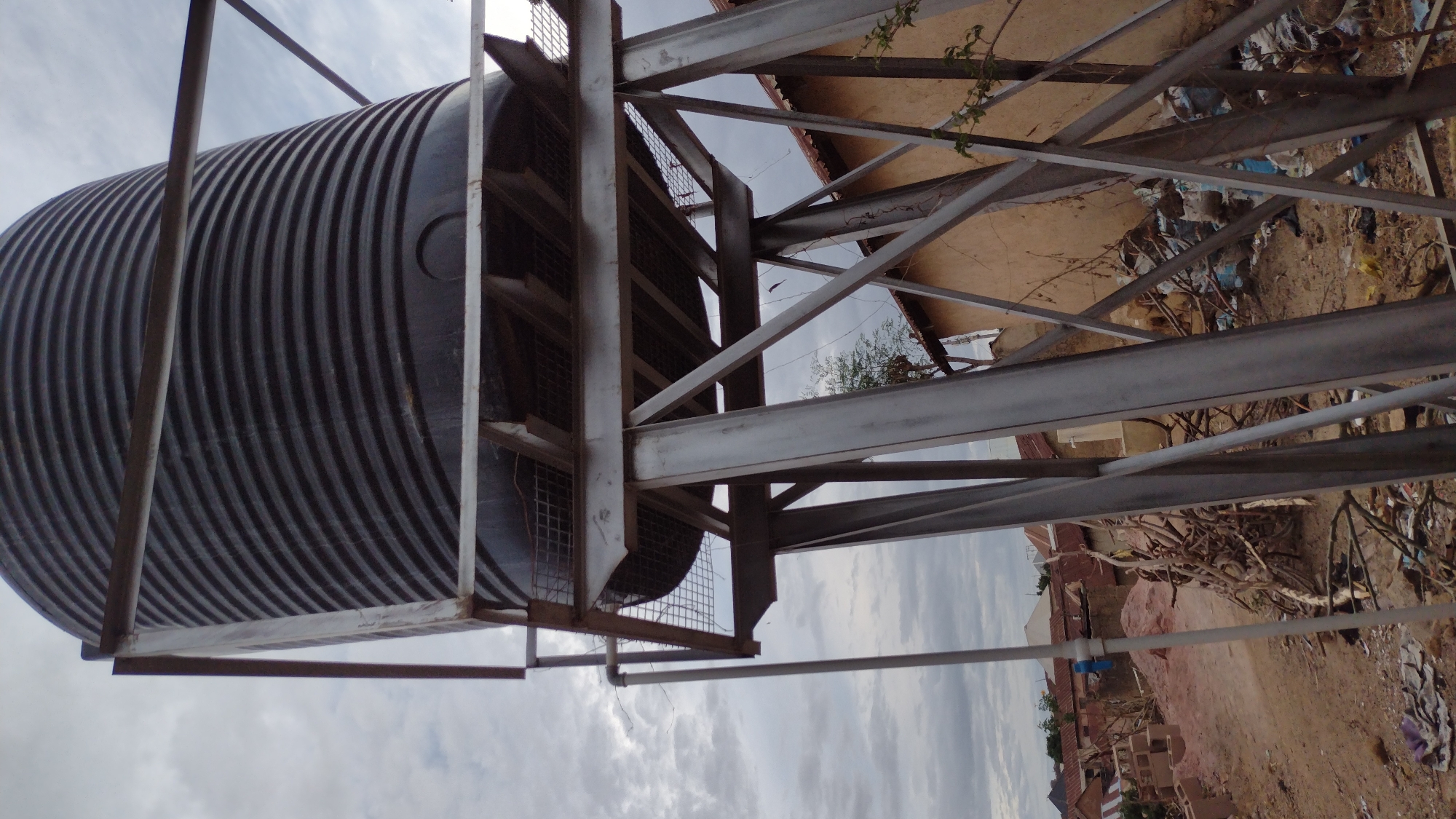 A large, black, corrugated water tank is mounted on a gray, steel frame. The frame is constructed of intersecting beams and a wire mesh platform. In the background, there are residential buildings with reddish-brown roofs, along with scattered trash and vegetation. The sky is overcast. The scene appears to be in an urban or semi-urban environment in Jos, Nigeria.